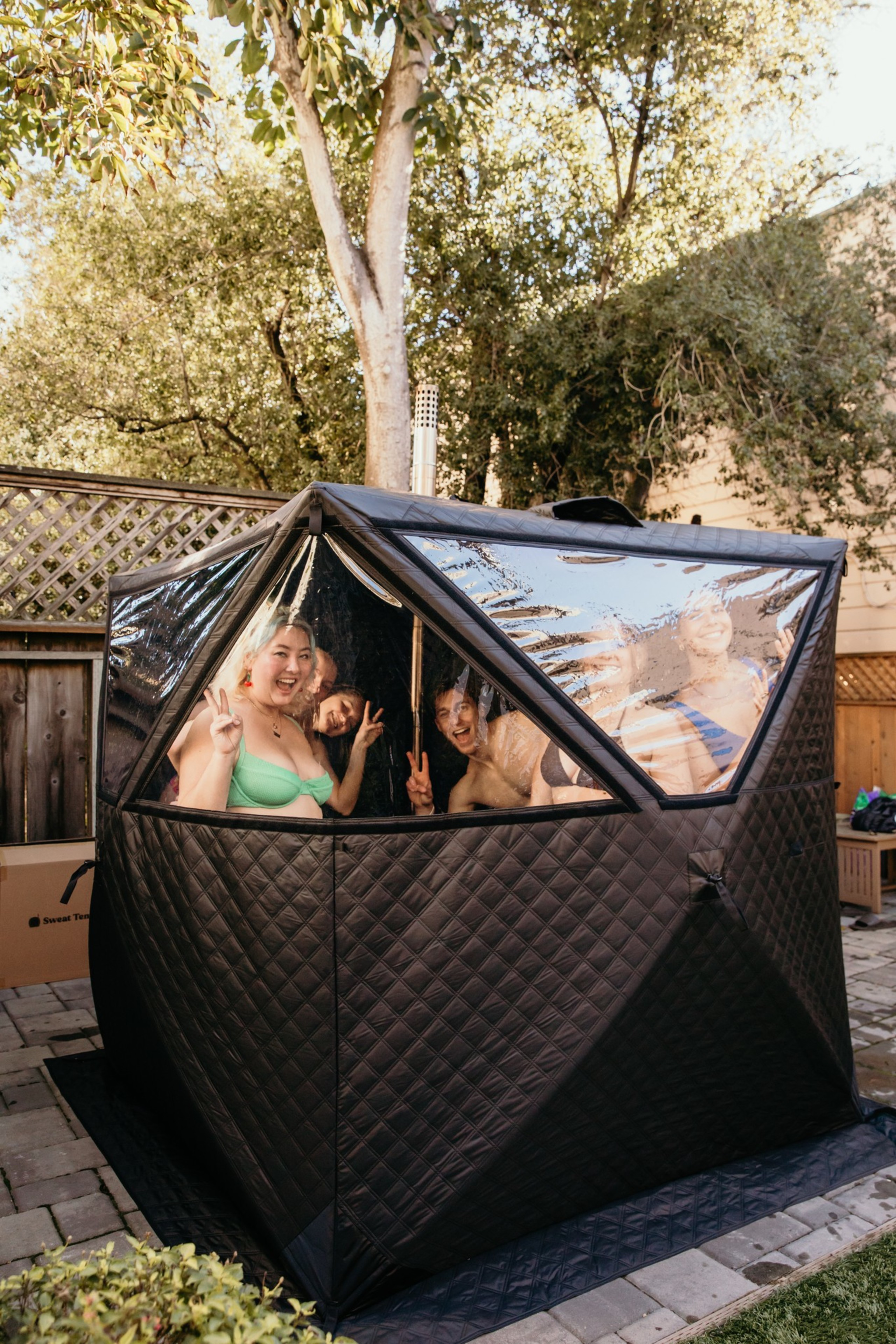 A group of people are inside a black, cube-shaped tent with transparent panels, smiling and making peace signs. The tent is set up outdoors with trees in the background.