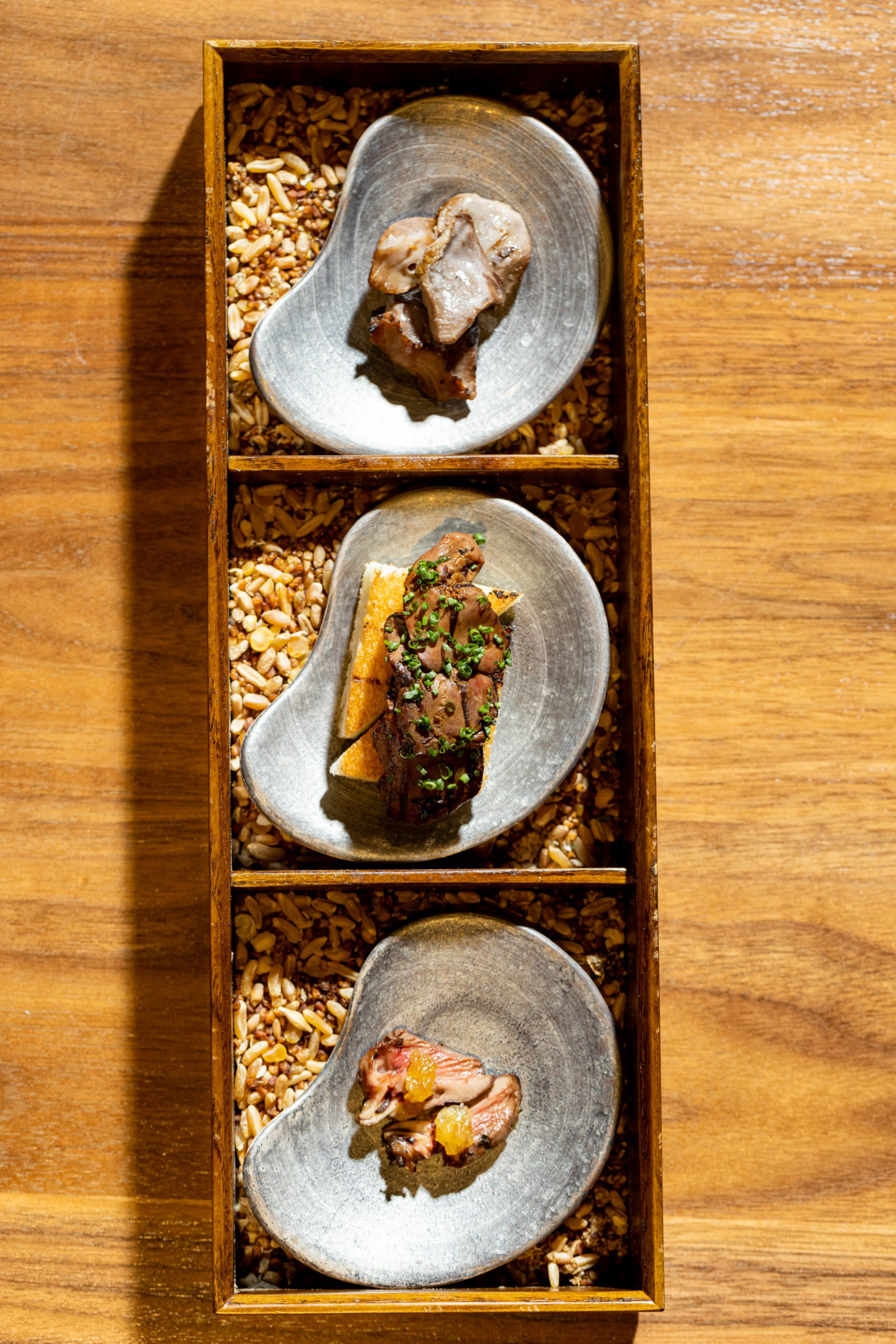 Three small portions of gourmet food served on curved silver plates, placed in a wooden box filled with grains, set on a wooden surface.