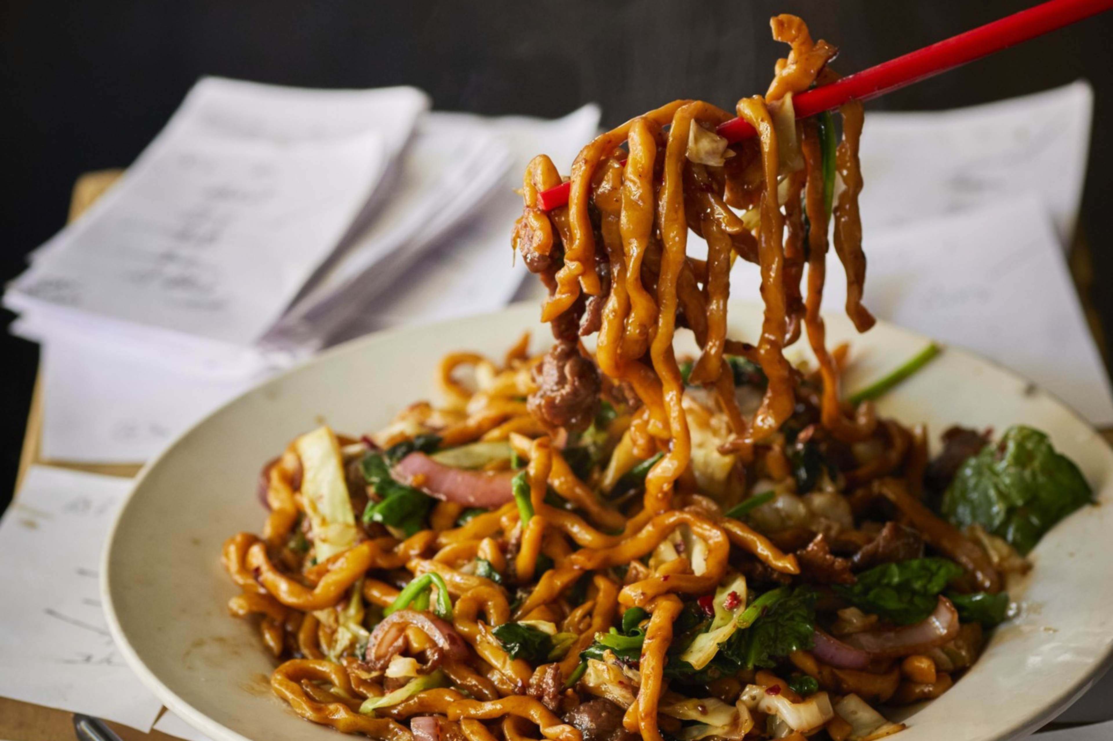 A plate of stir-fried noodles with vegetables and meat is being lifted by red chopsticks, placed on a cardboard box covered with handwritten notes and pens.