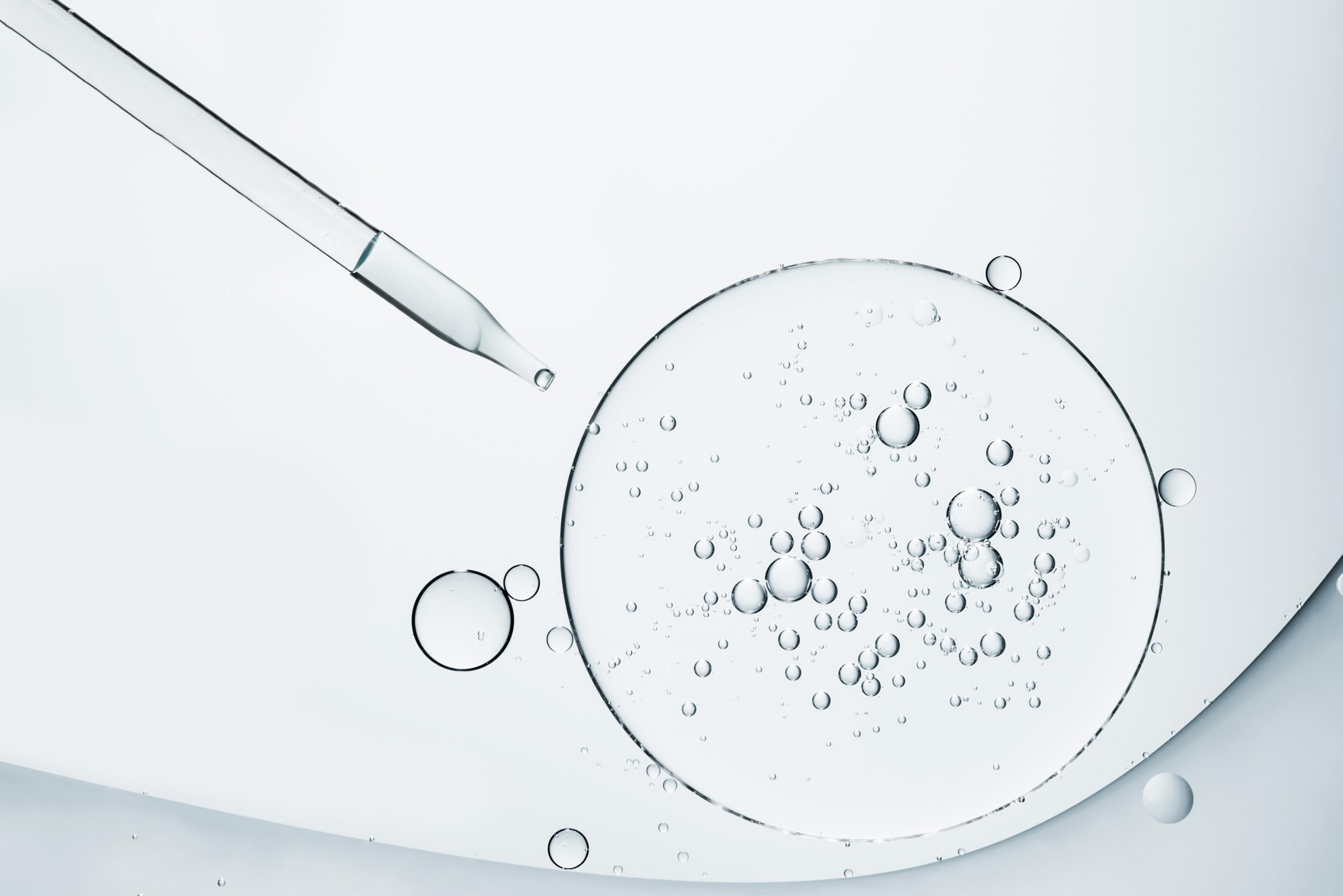 The image shows a clear liquid-filled dropper above large circular surfaces containing bubbles on a smooth, light background.