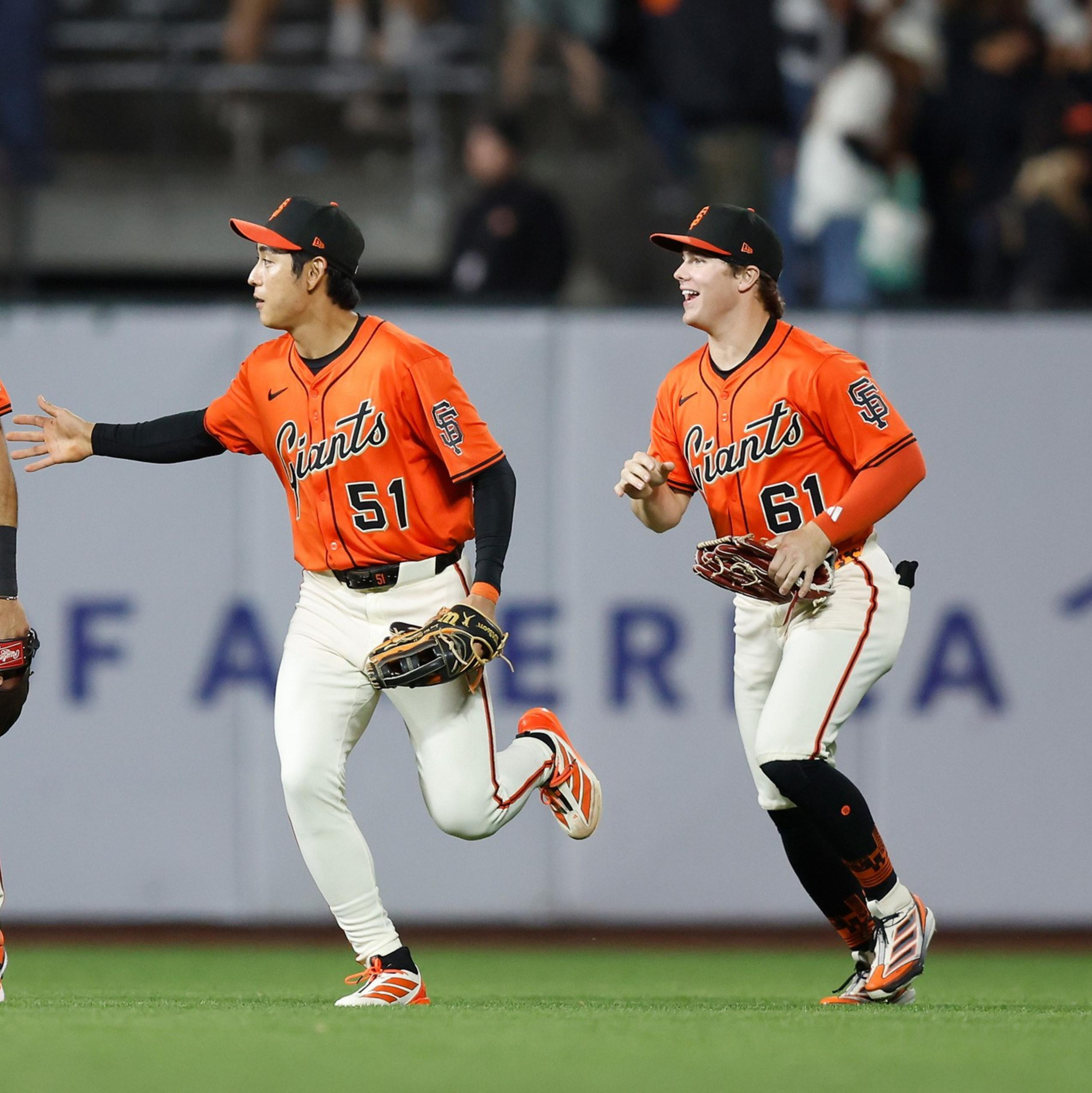 Three baseball players in orange and white Giants uniforms celebrate together on the field, smiling and running toward each other.