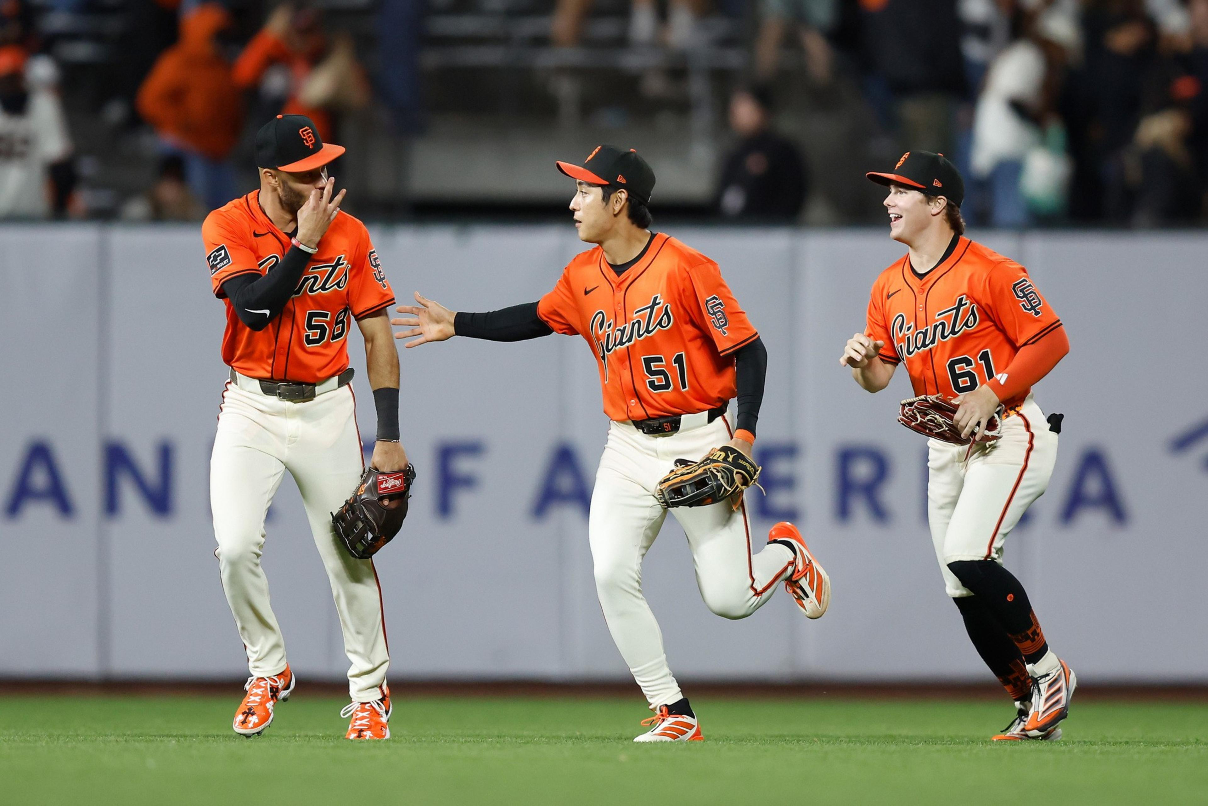 Three baseball players in orange and white Giants uniforms celebrate together on the field, smiling and running toward each other.