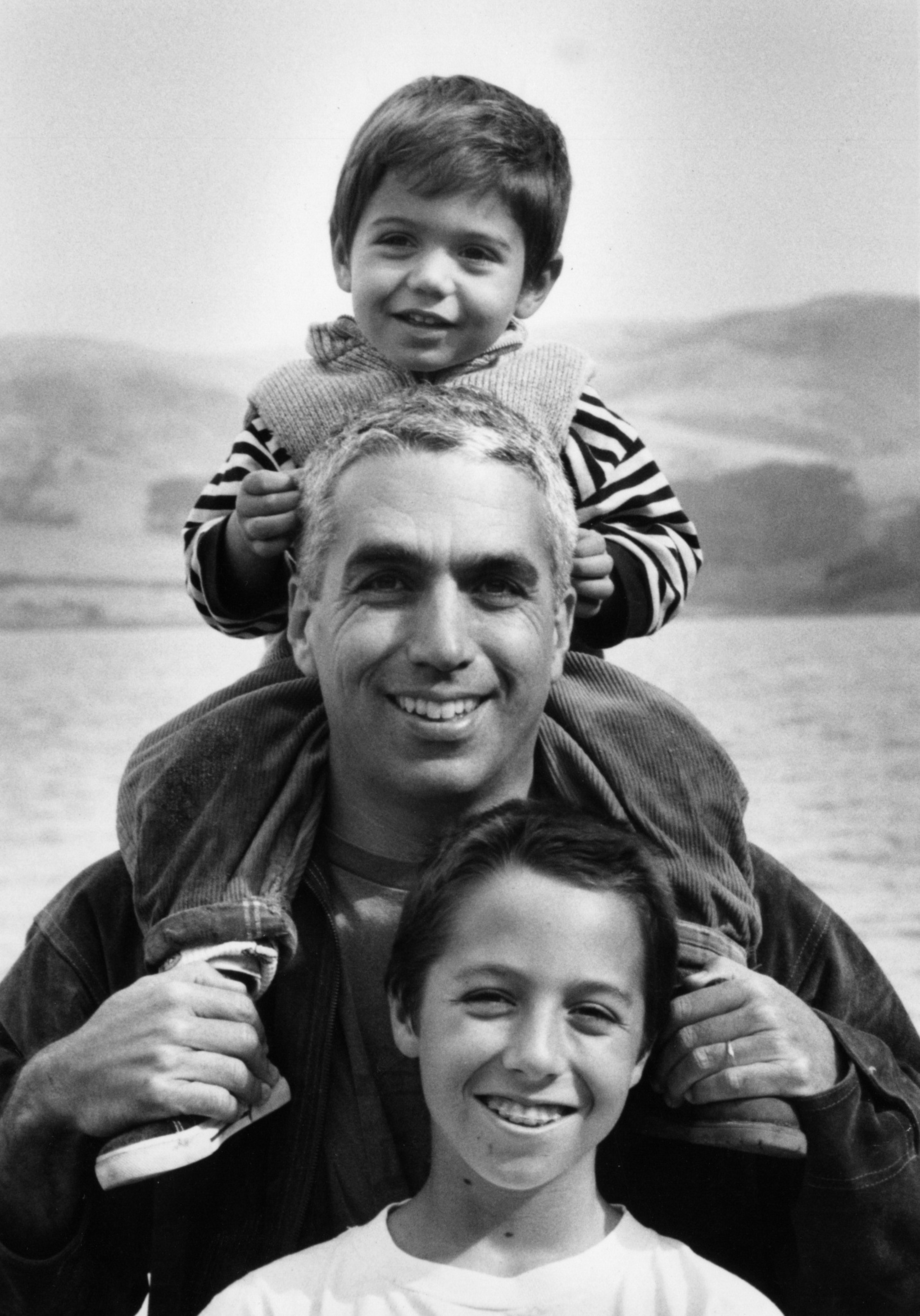 A man smiles while holding a child on his shoulders. Another child stands in front, also smiling. The background shows a calm lake and hills.