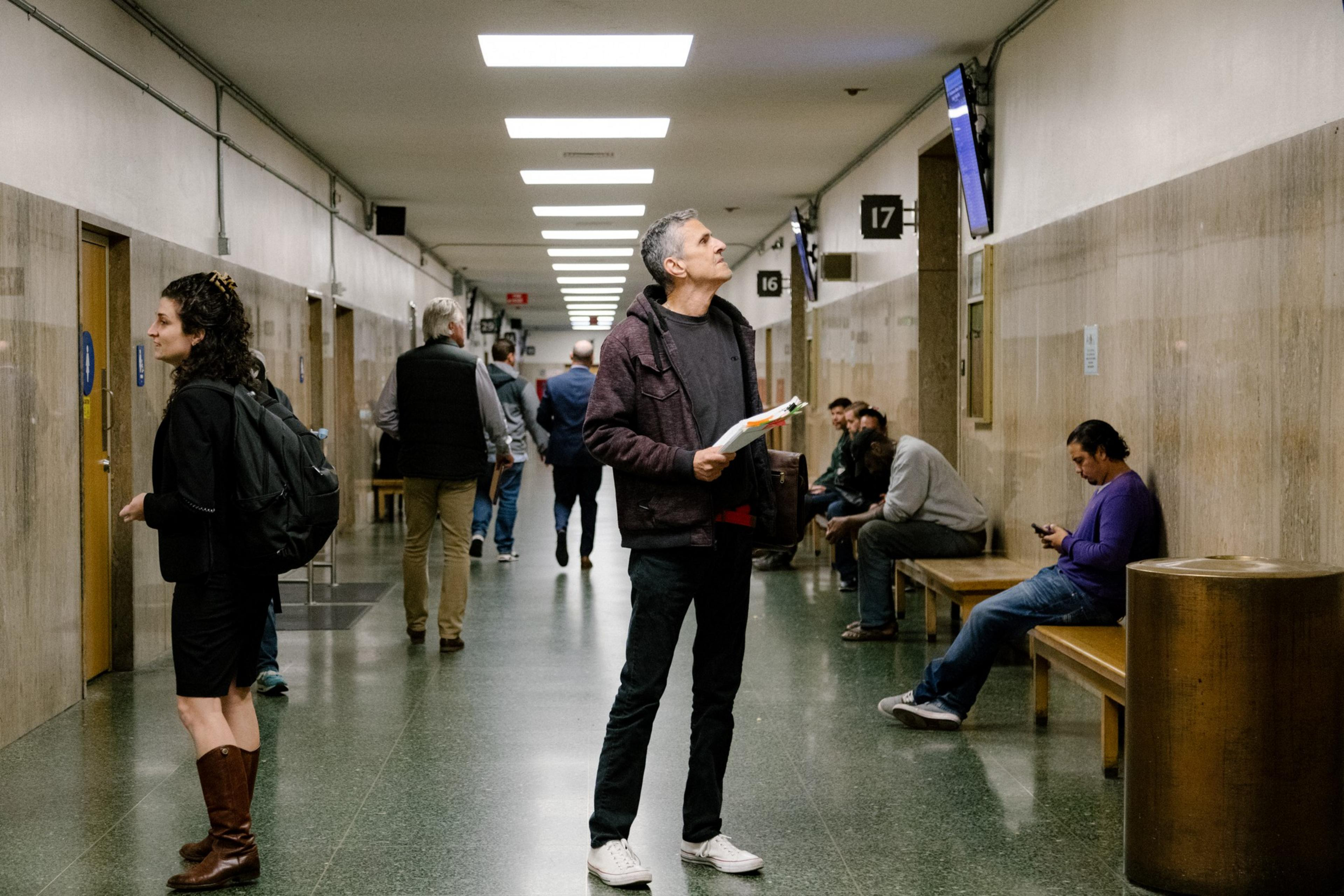 A man holds documents while looking up a monitor in a long courtroom hallway as other people walk through the space.