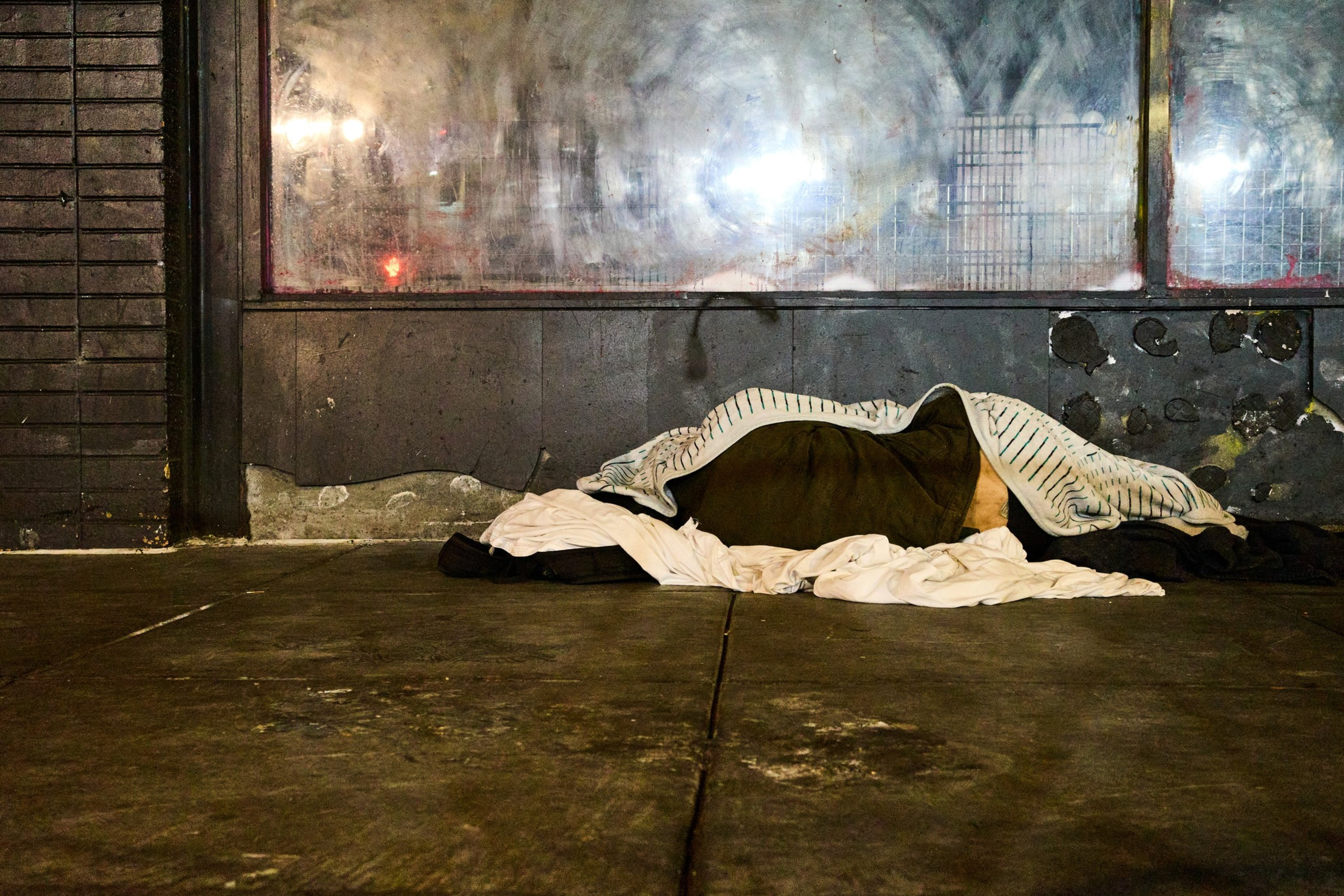 A person is sleeping on the ground under a blanket in an urban setting. The backdrop is a weathered, dirty wall with a window reflecting lights.
