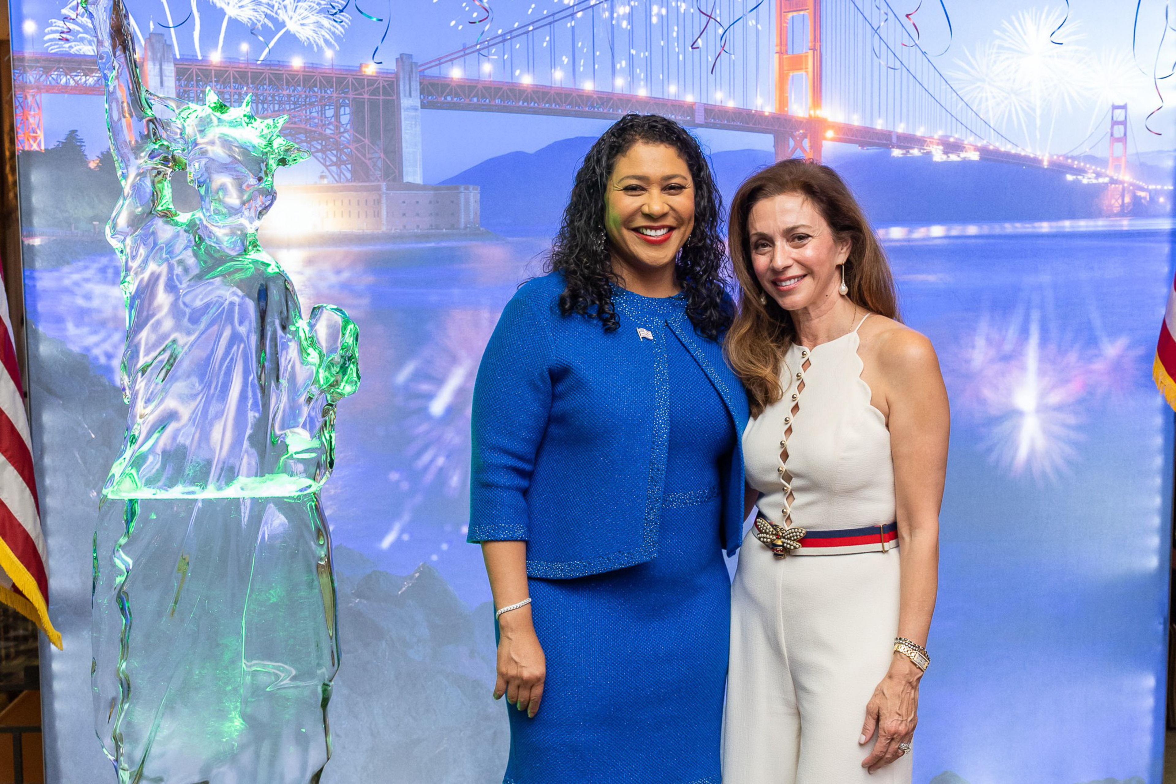 Two women smile and pose in front of a backdrop of the Golden Gate Bridge at night with fireworks, next to an ice sculpture of the Statue of Liberty.