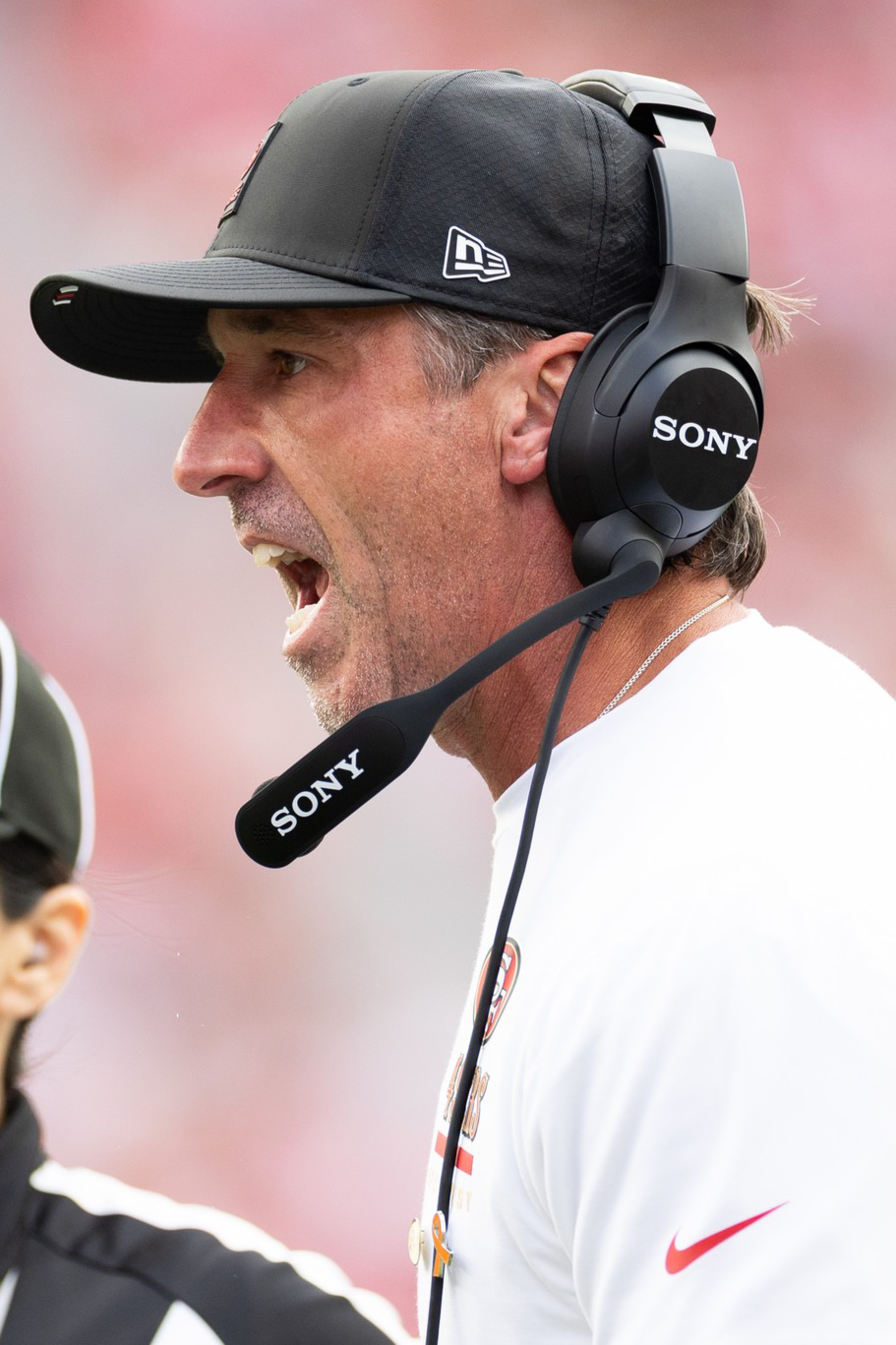 A football coach wearing a black cap and headset is shouting, while a female referee with an NFL logo hat listens attentively nearby.