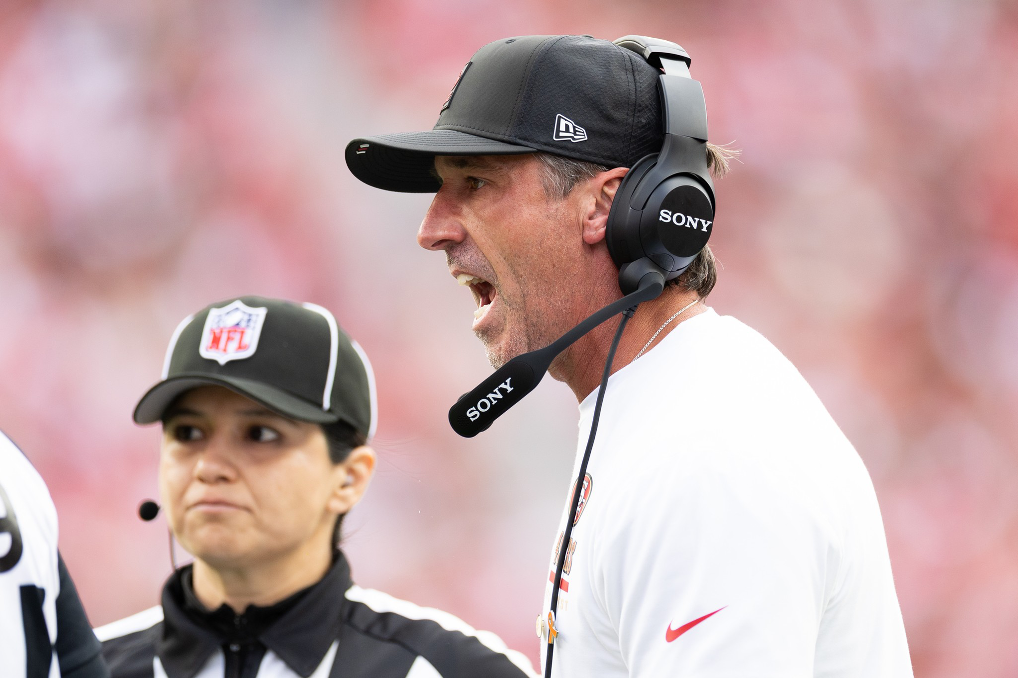 A football coach wearing a black cap and headset is shouting, while a female referee with an NFL logo hat listens attentively nearby.