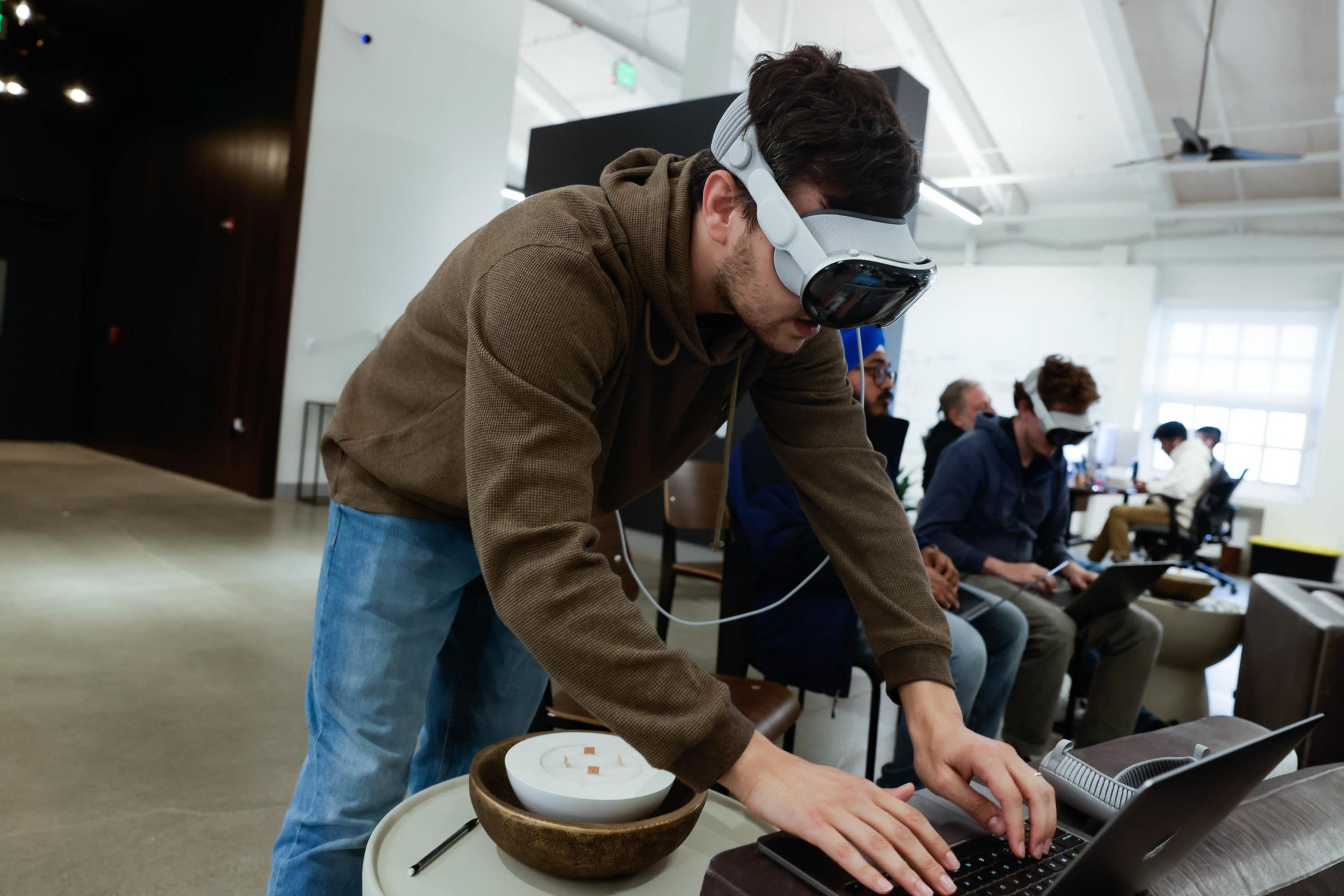 A person in a hoodie and Apple Vision Pro headset leans over a laptop and types in a busy office-like setting.