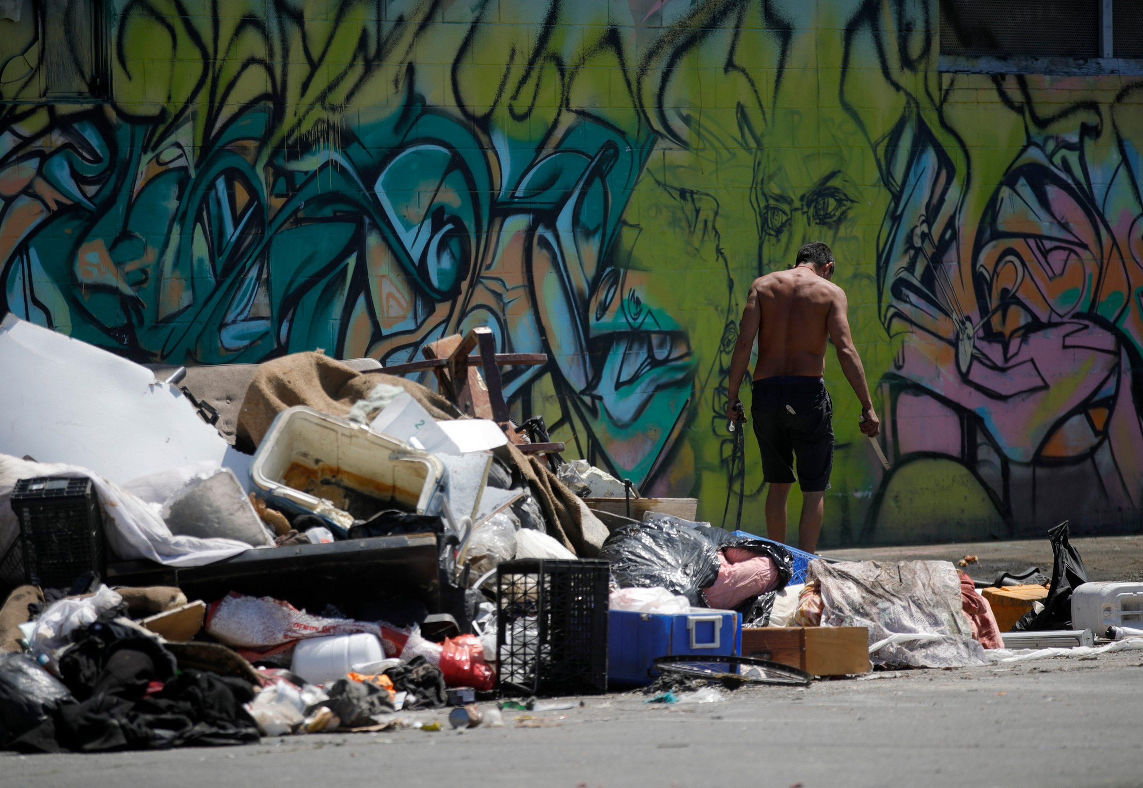 A shirtless man stands near a pile of trash in front of a colorful graffiti-covered wall on a sunny day.
