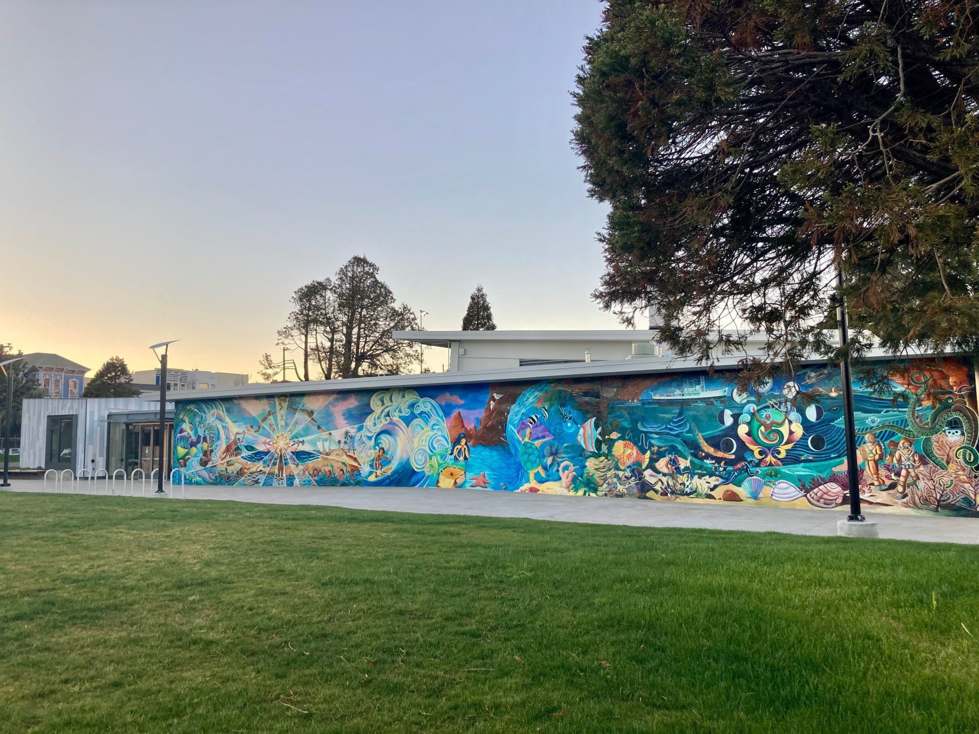 A colorful mural on a long wall depicts ocean waves, marine life, seashells, and underwater scenes blending fantasy and nature beside a grassy area.