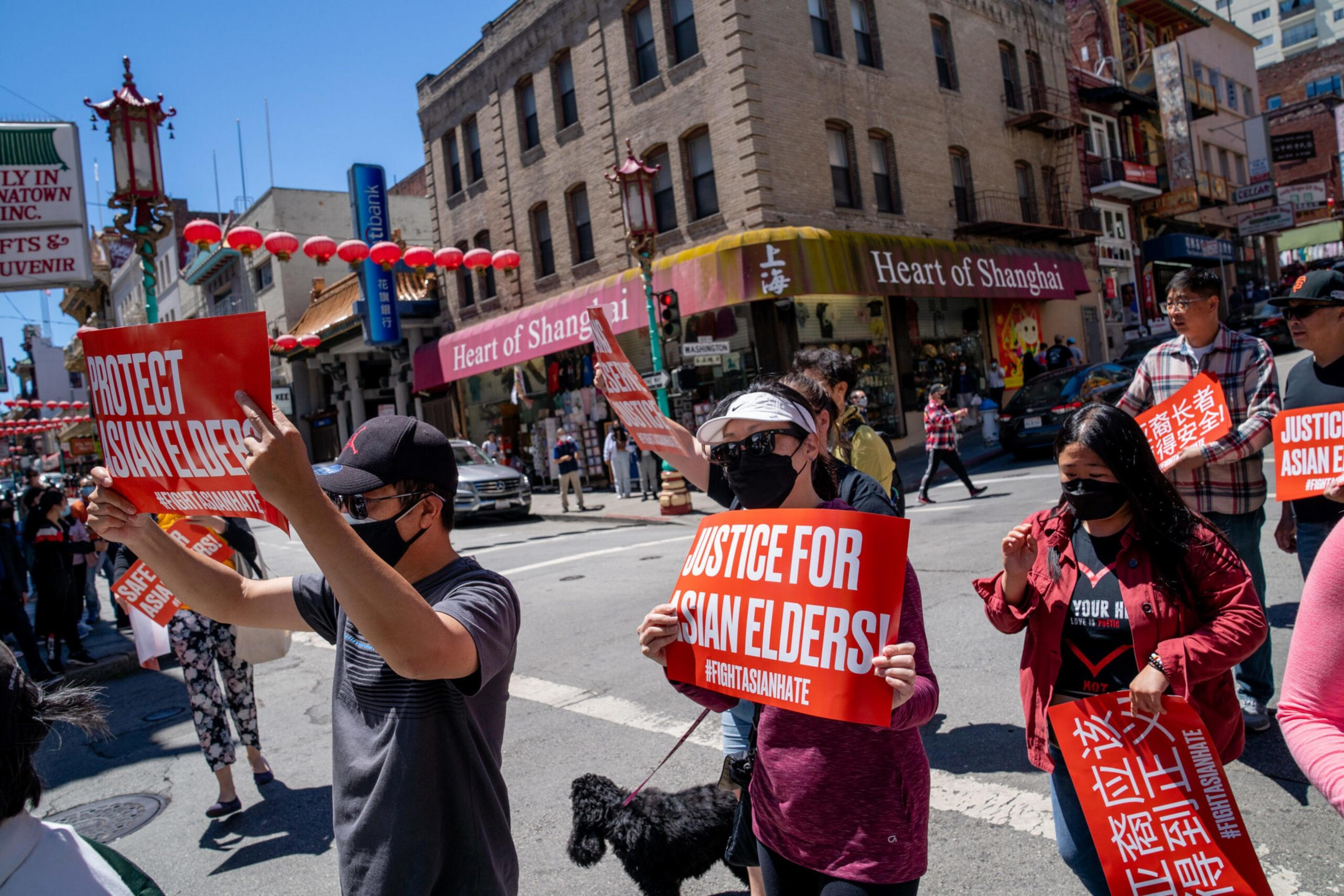 People wearing masks hold red signs reading "Protect Asian Elders" and "Justice for Asian Elders" while walking in a busy Chinatown street.