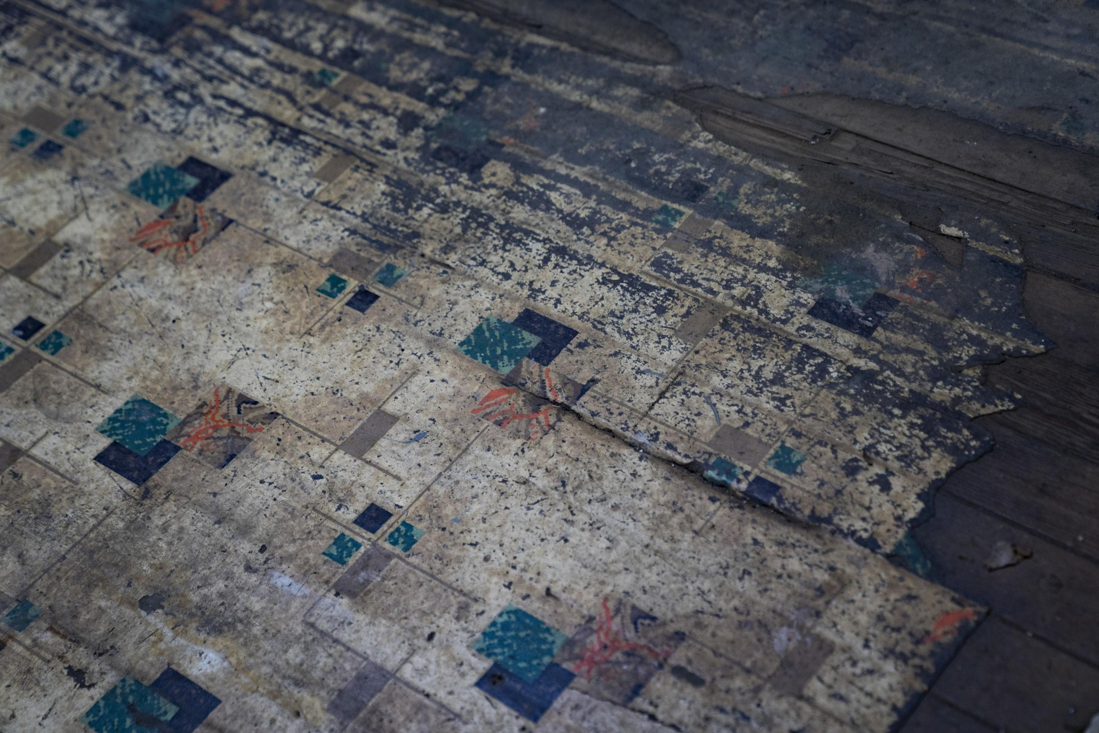This image shows an old, worn-out linoleum floor with a geometric pattern of squares, some in green and black. The edges are peeling, revealing wood underneath.