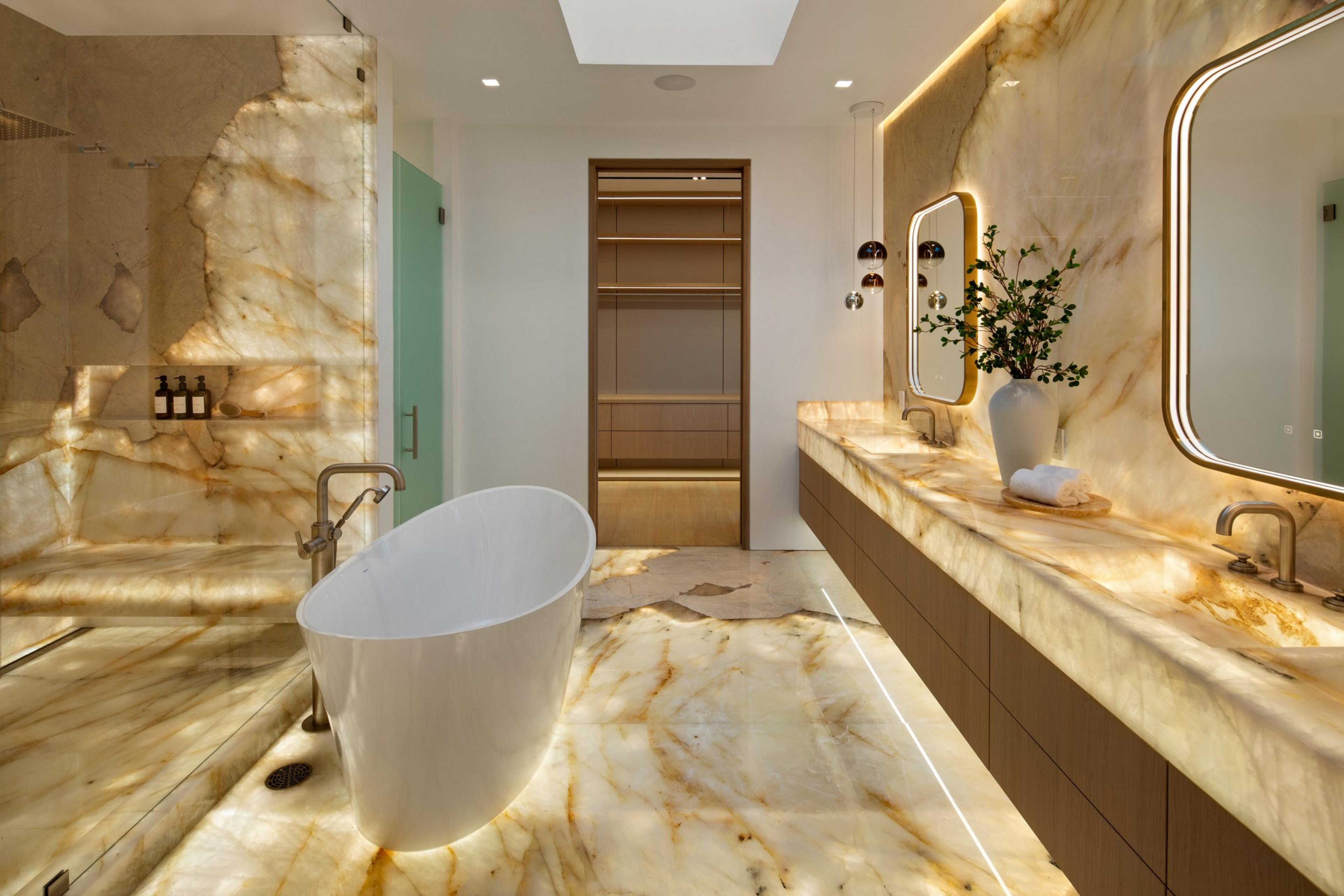 A luxurious bathroom features a freestanding white tub, illuminated marble floors and walls, double sinks with backlit mirrors, and a glass shower with built-in shelves.