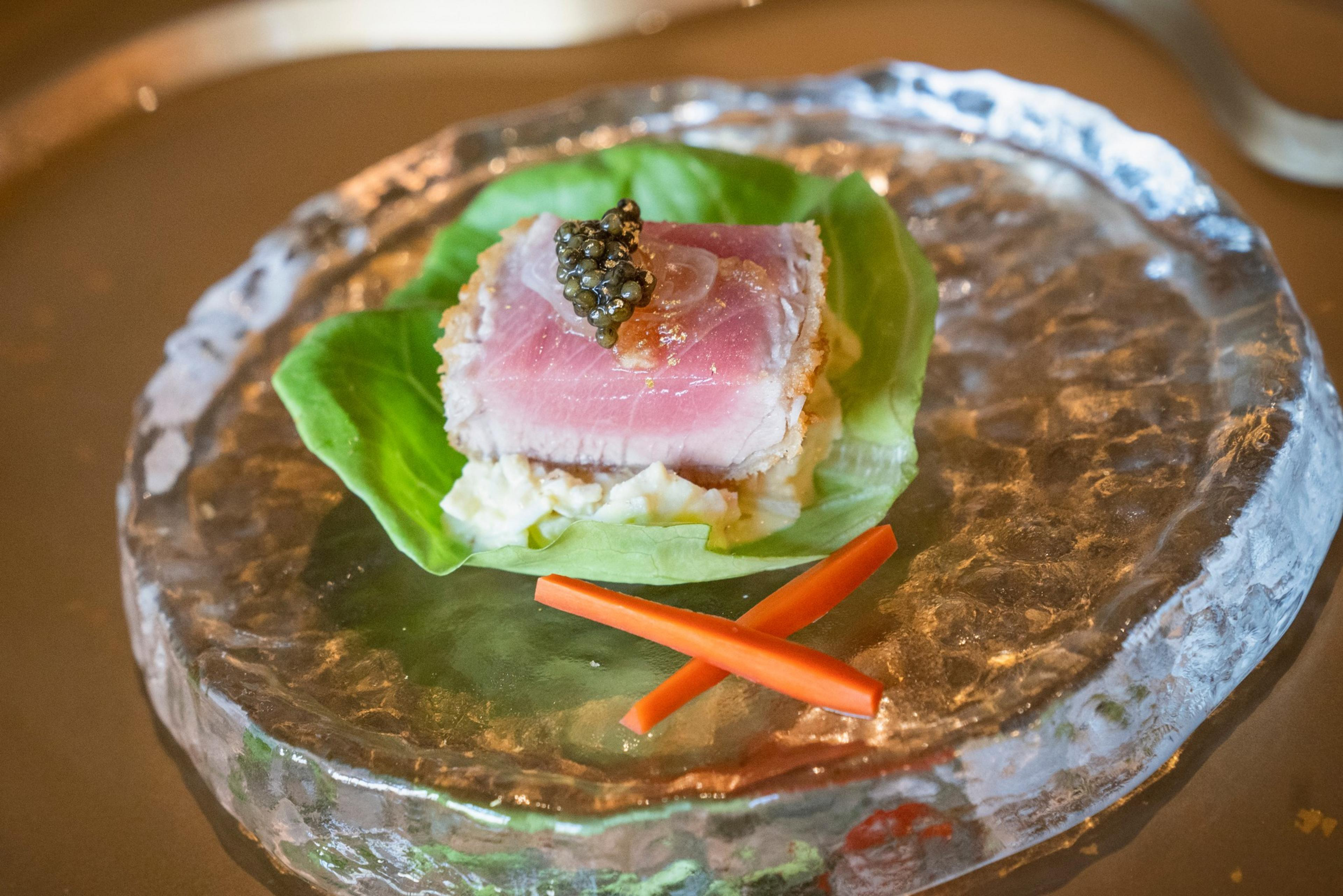 A slice of seared tuna topped with caviar sits on a lettuce leaf with a creamy base, garnished with two thin carrot sticks, served on a textured glass plate.