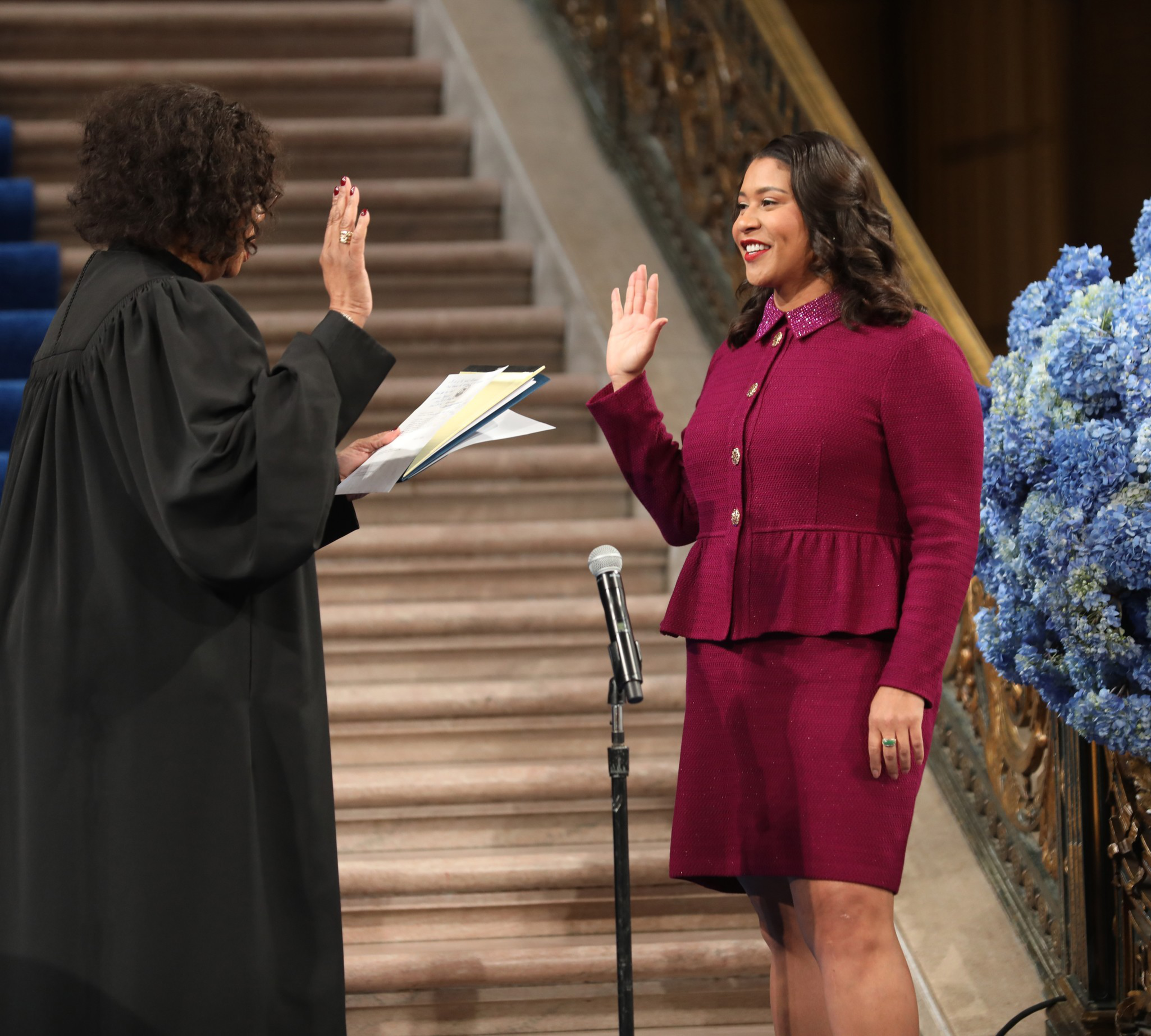A person in a judicial robe is administering an oath to a woman in a burgundy suit, both raising their right hands. A microphone and blue flowers are nearby.
