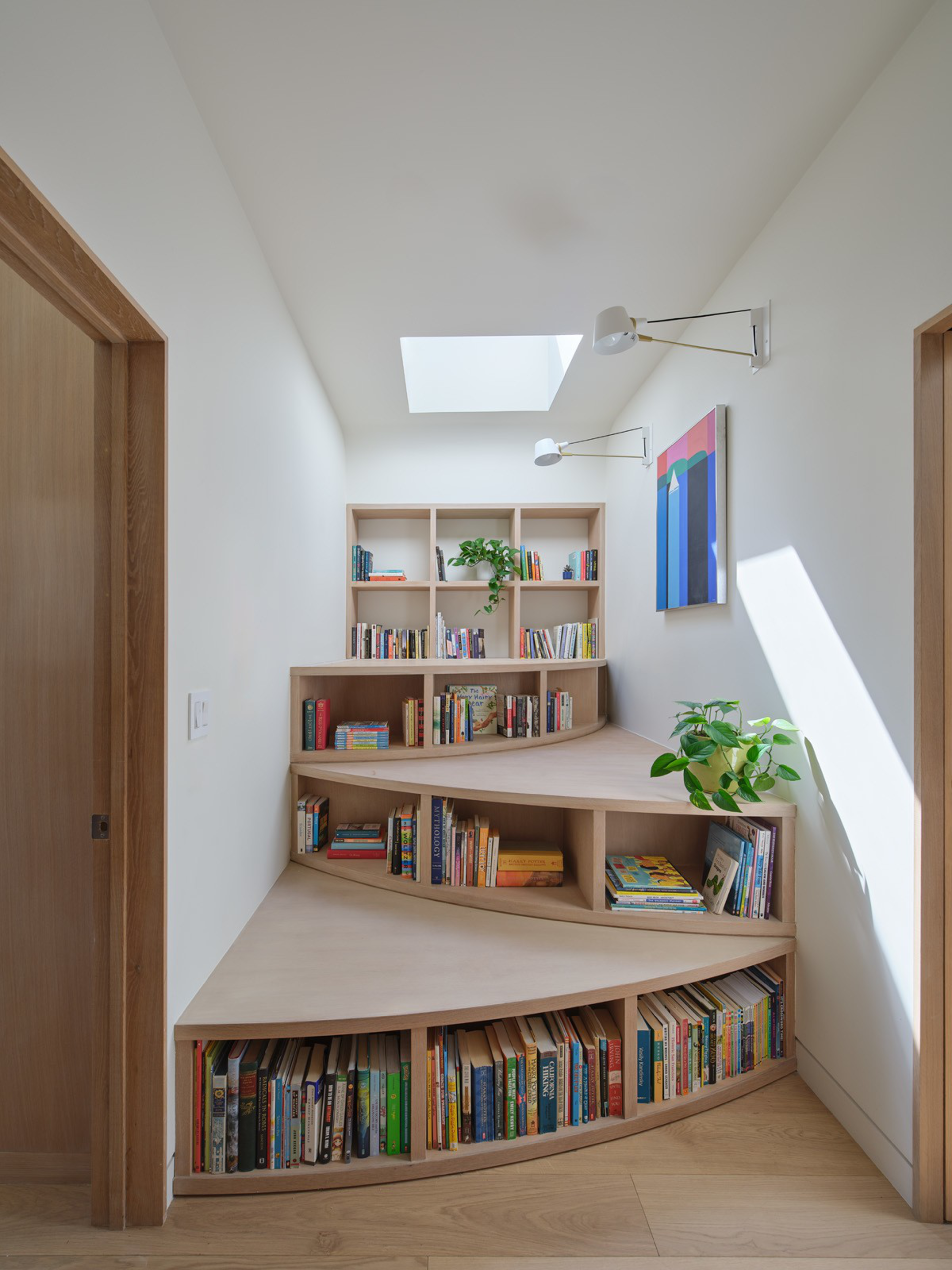 A built-in, curved wooden bookshelf filled with books and plants sits under a skylight in a narrow, white-walled space with a colorful wall painting and two wall lamps.