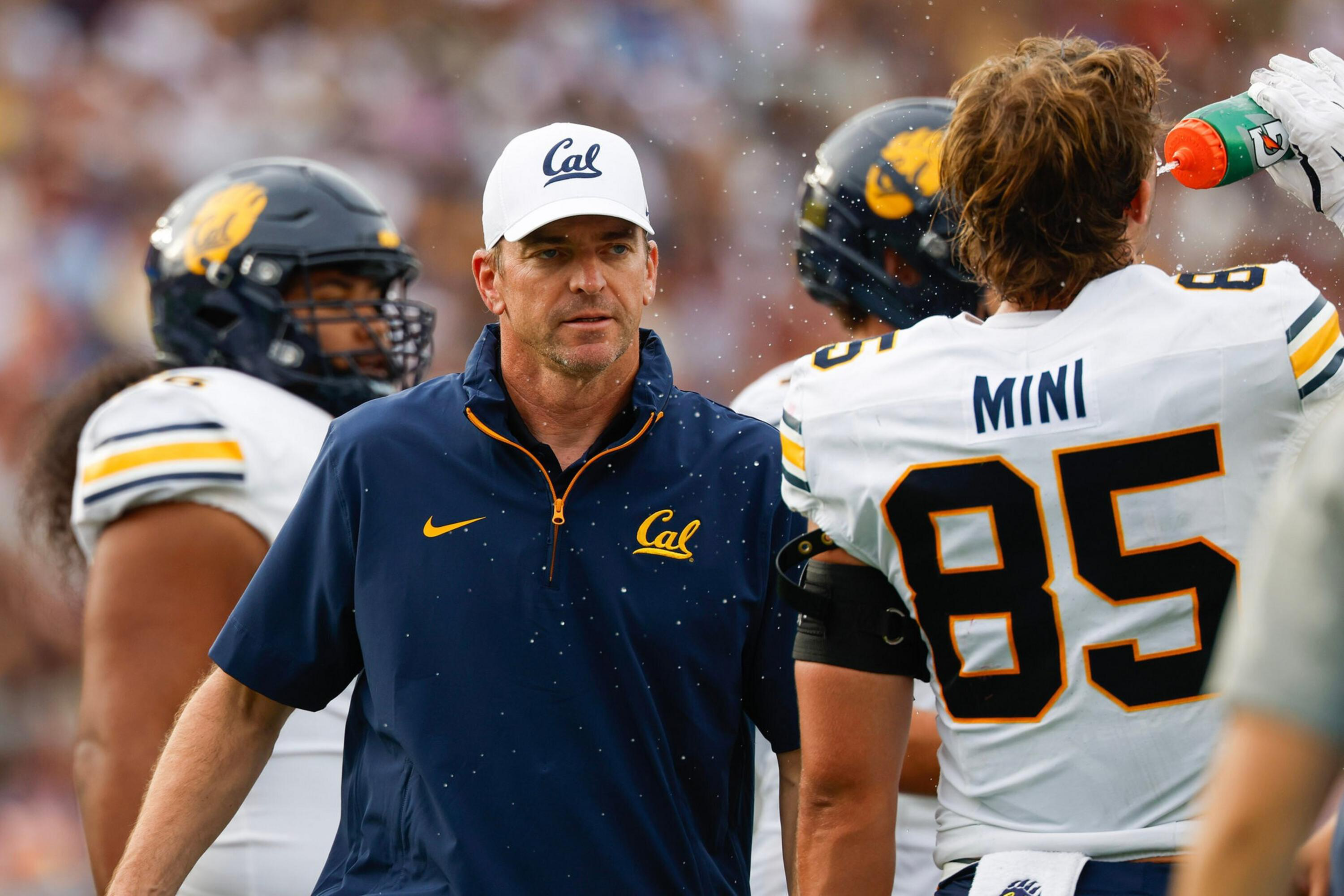 A football coach wearing a white Cal cap and navy Cal jacket stands near a player in a white jersey numbered 85, while another player drinks from a bottle.
