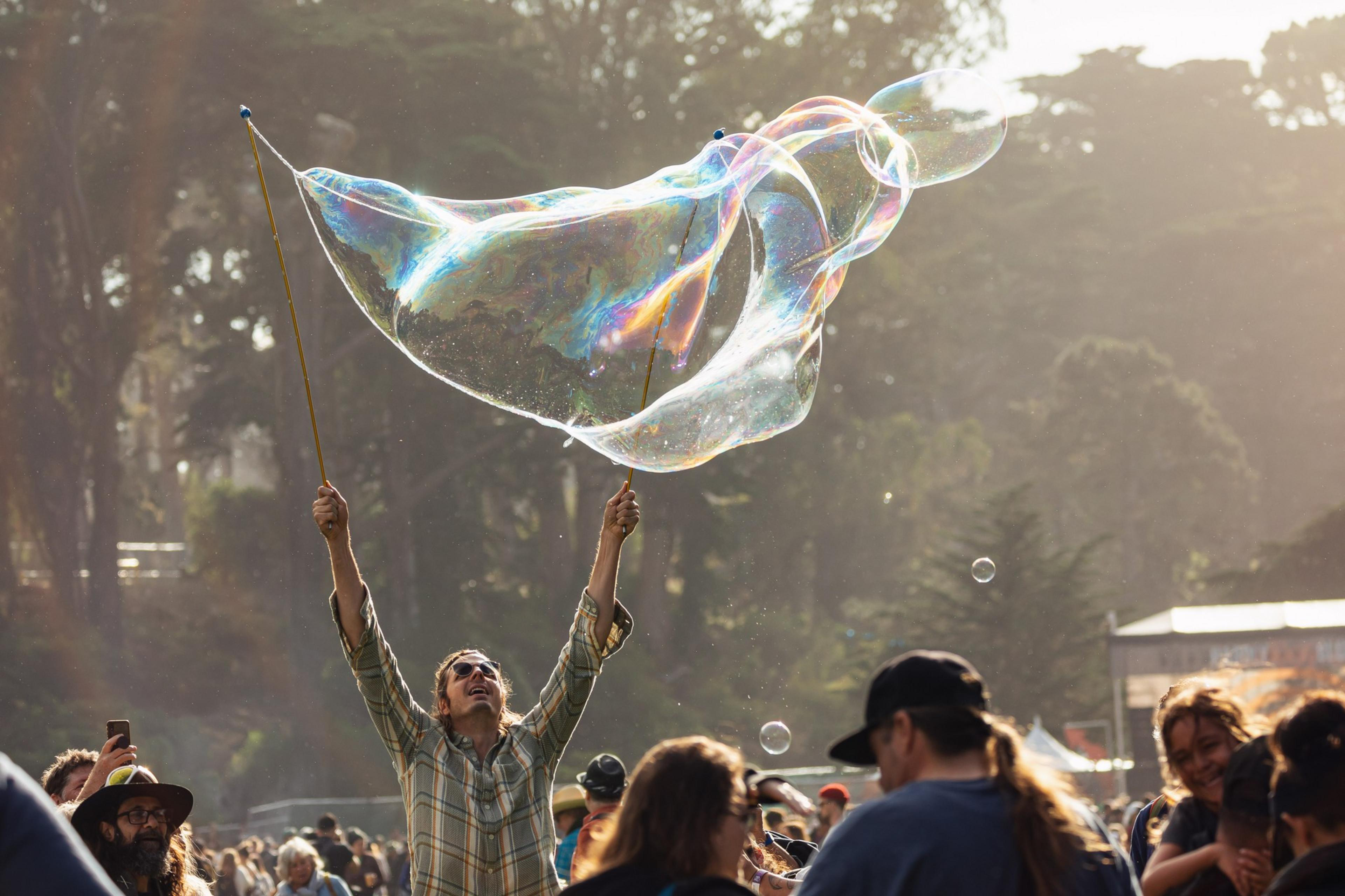 A person in a plaid shirt creates a large, colorful bubble using two sticks at an outdoor event, surrounded by a crowd with trees in the background.