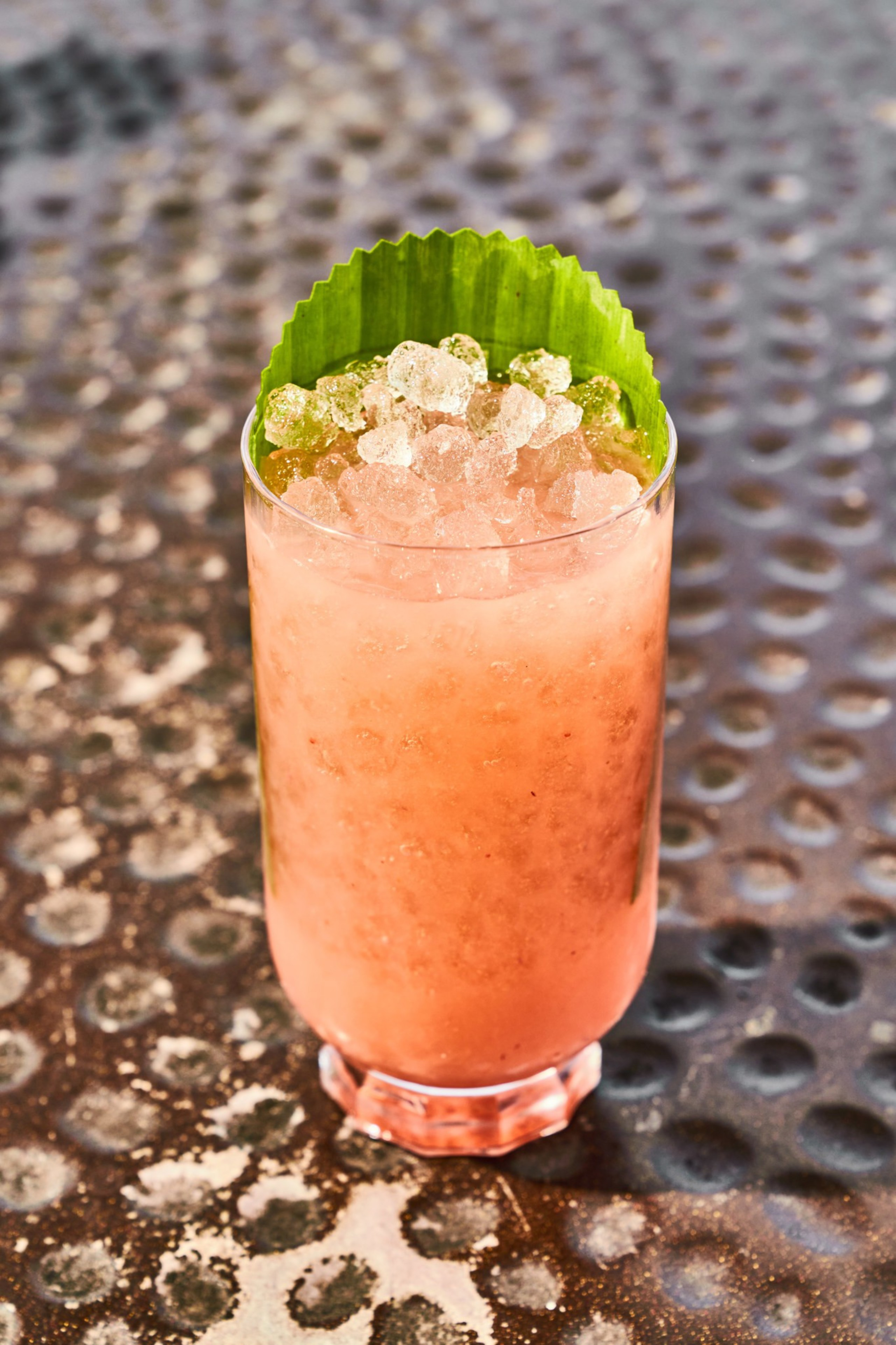 A pink slushy drink in a glass is topped with crushed ice and a green leaf, resting on a textured surface.