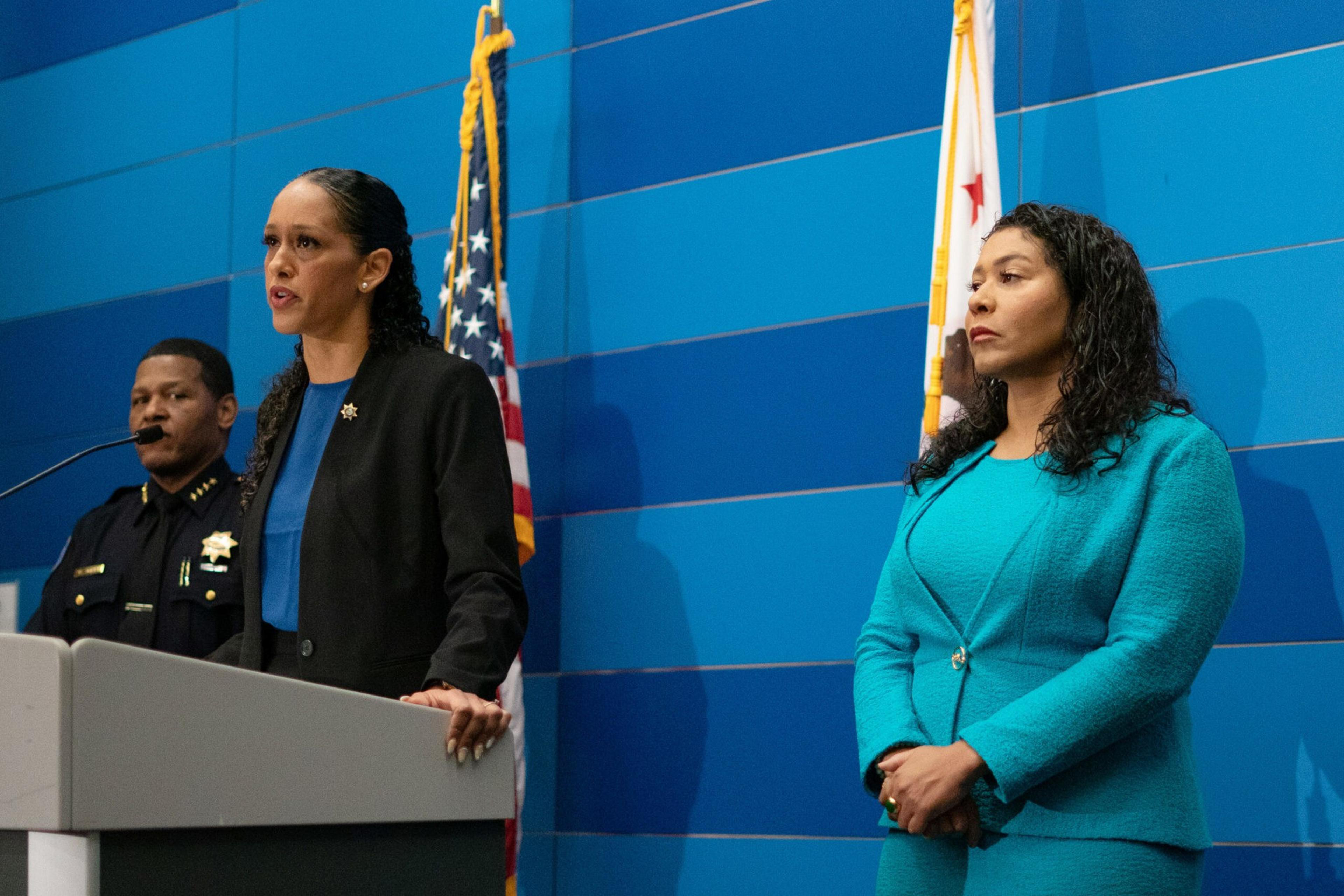 A woman in a black blazer speaks at a podium with two flags behind her, while a man in a police uniform and another woman in a turquoise suit stand nearby.
