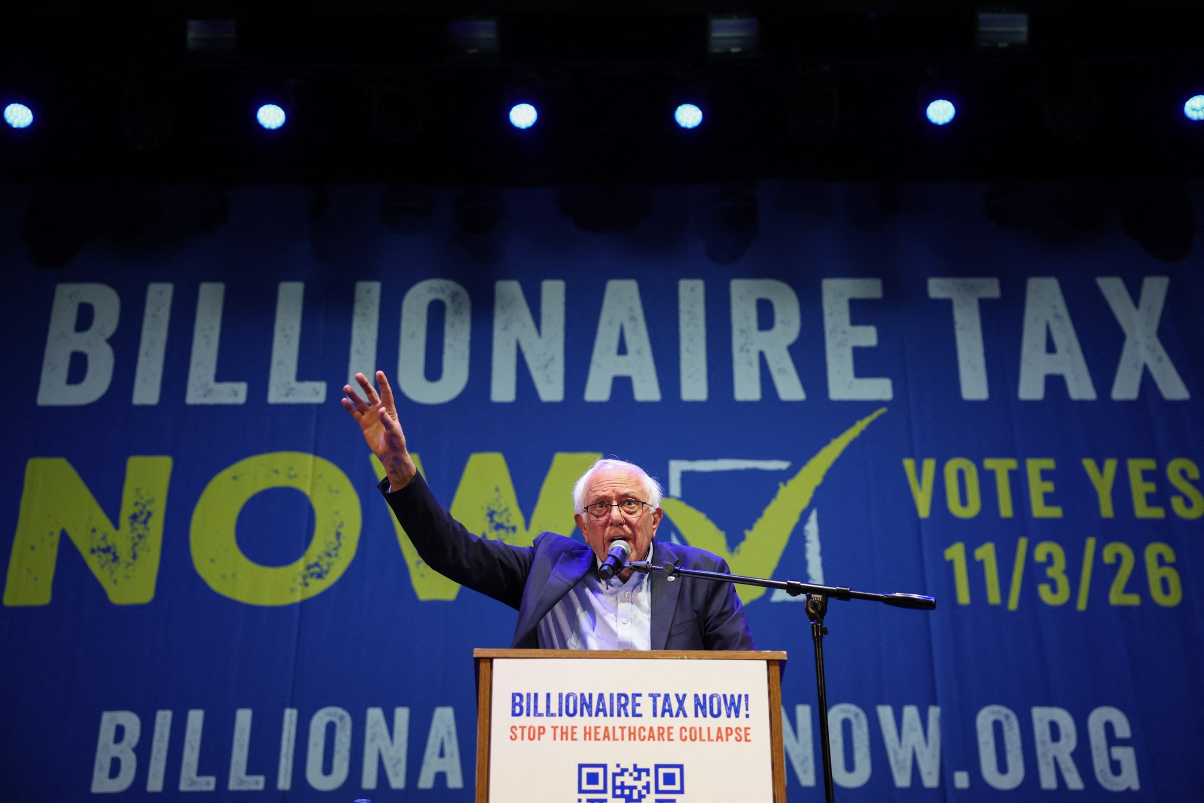 A man speaks passionately at a podium with a microphone, raising his hand in front of a large banner that says "Billionaire Tax Now" and voting information.