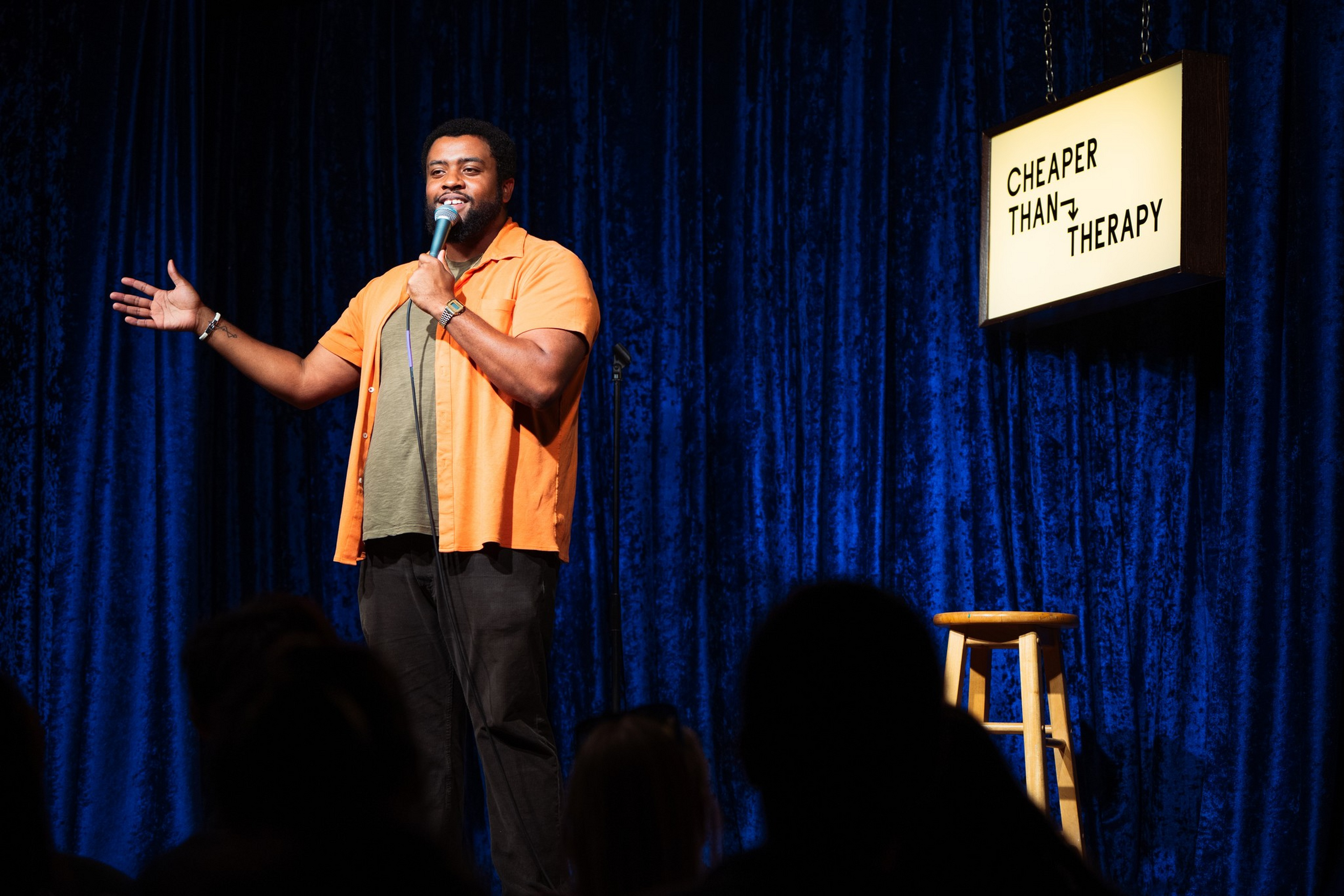 A man in an orange shirt holds a microphone, performing comedy on stage with a stool nearby and a sign reading “CHEAPER THAN THERAPY.”