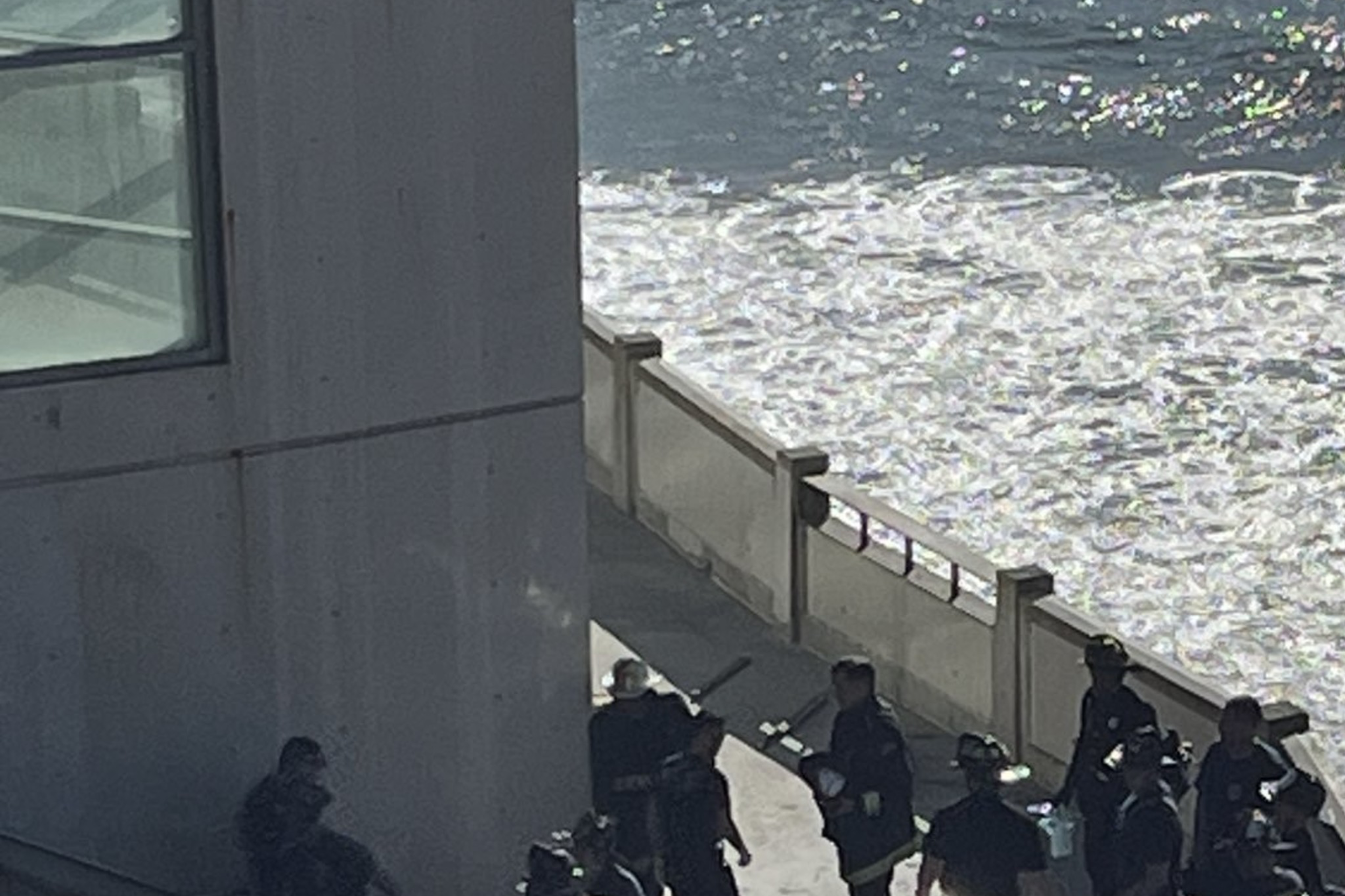 The image shows a car crash scene by the seaside, with emergency personnel gathered around a badly damaged vehicle at the base of a concrete structure.
