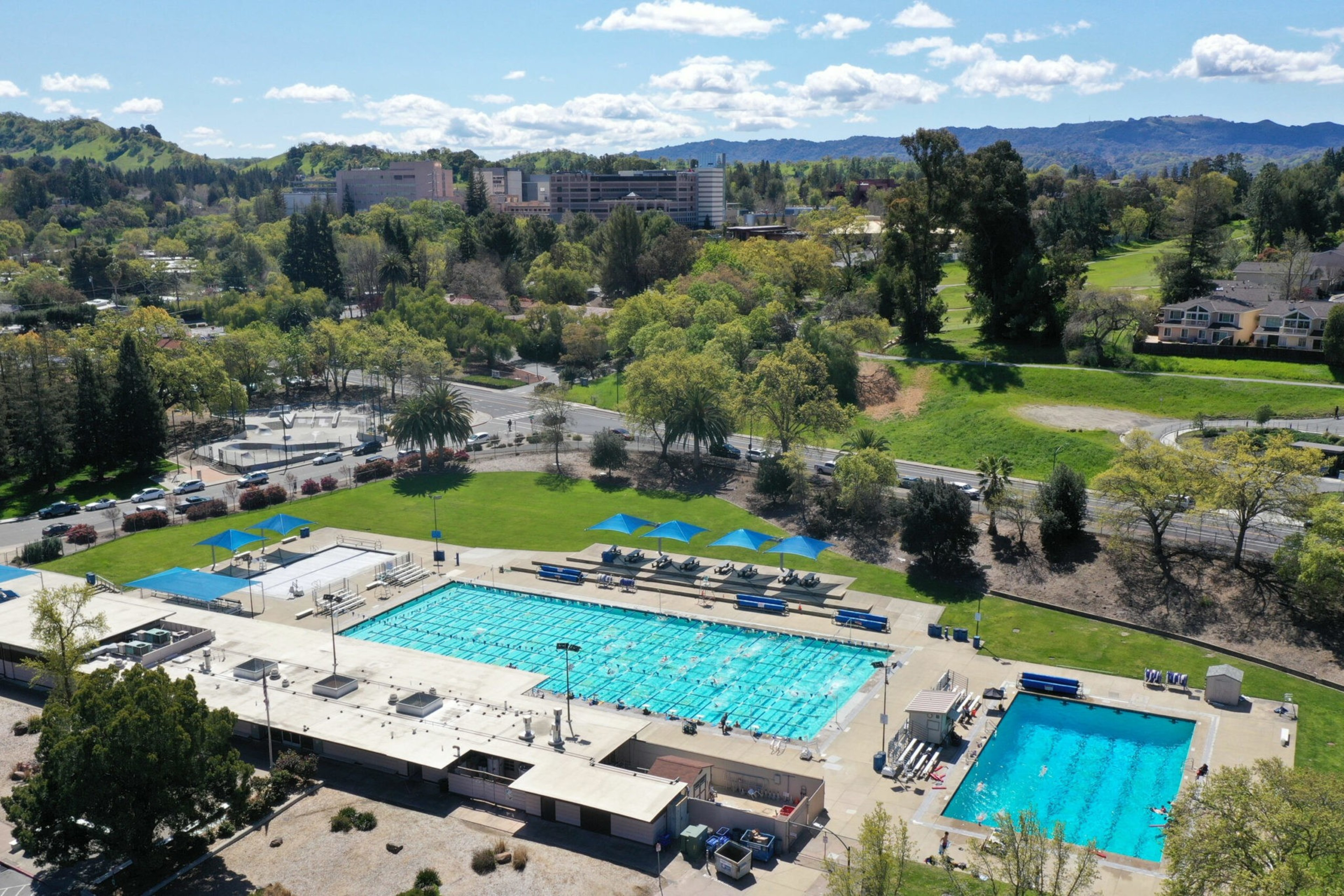 Two large outdoor swimming pools with swimmers, surrounded by benches, blue shade canopies, green lawns, trees, and distant hills under a blue sky.