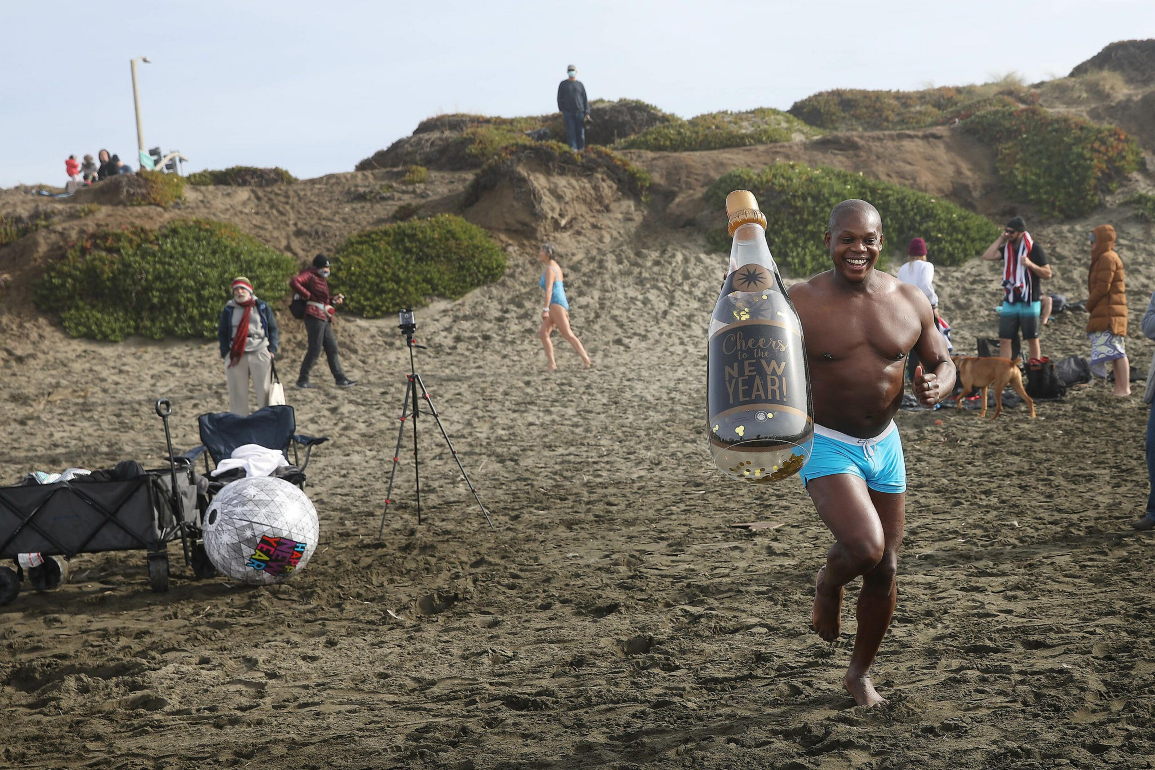 A joyful man in blue shorts runs barefoot on a sandy beach holding a large inflatable champagne bottle that says "Cheers to the New Year!"