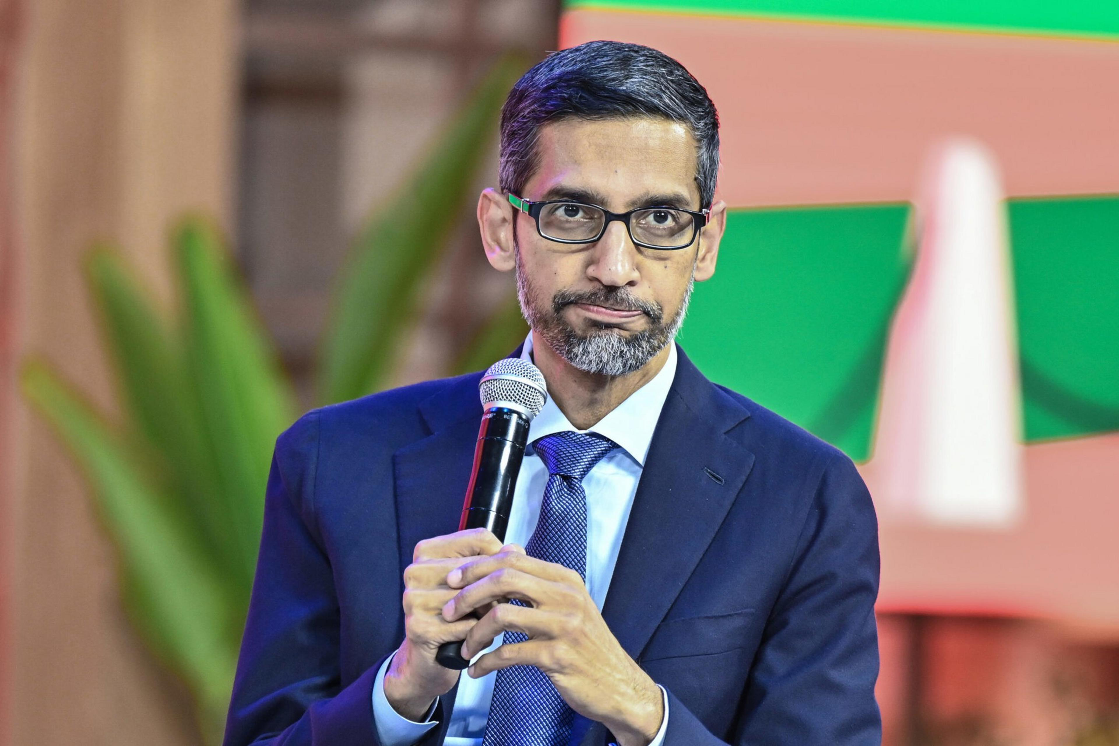 A man with glasses, a beard, and a mustache wears a blue suit and tie while holding a microphone and looking thoughtfully ahead.
