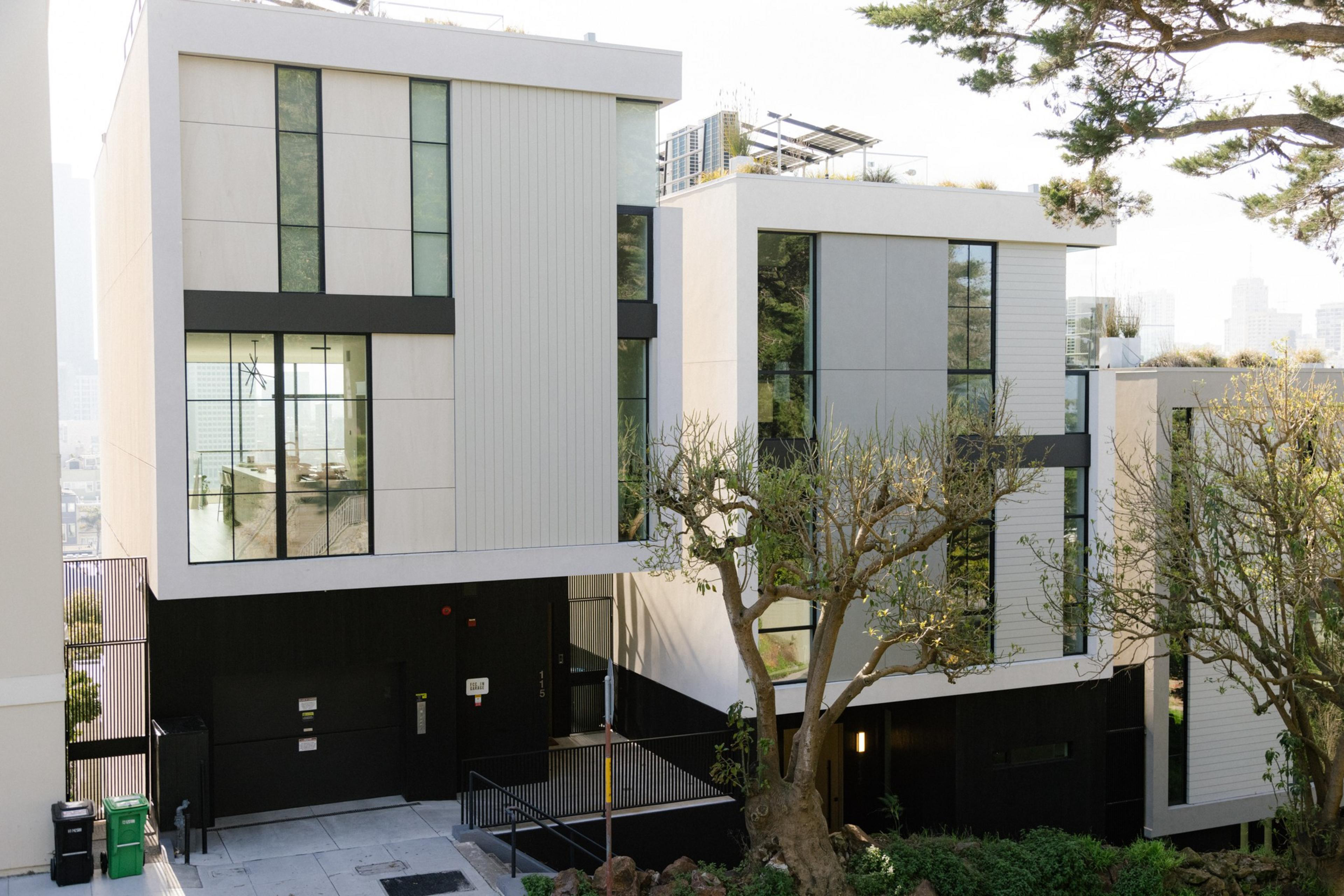 Modern buildings with large windows amid trees; city skyline in the distance.