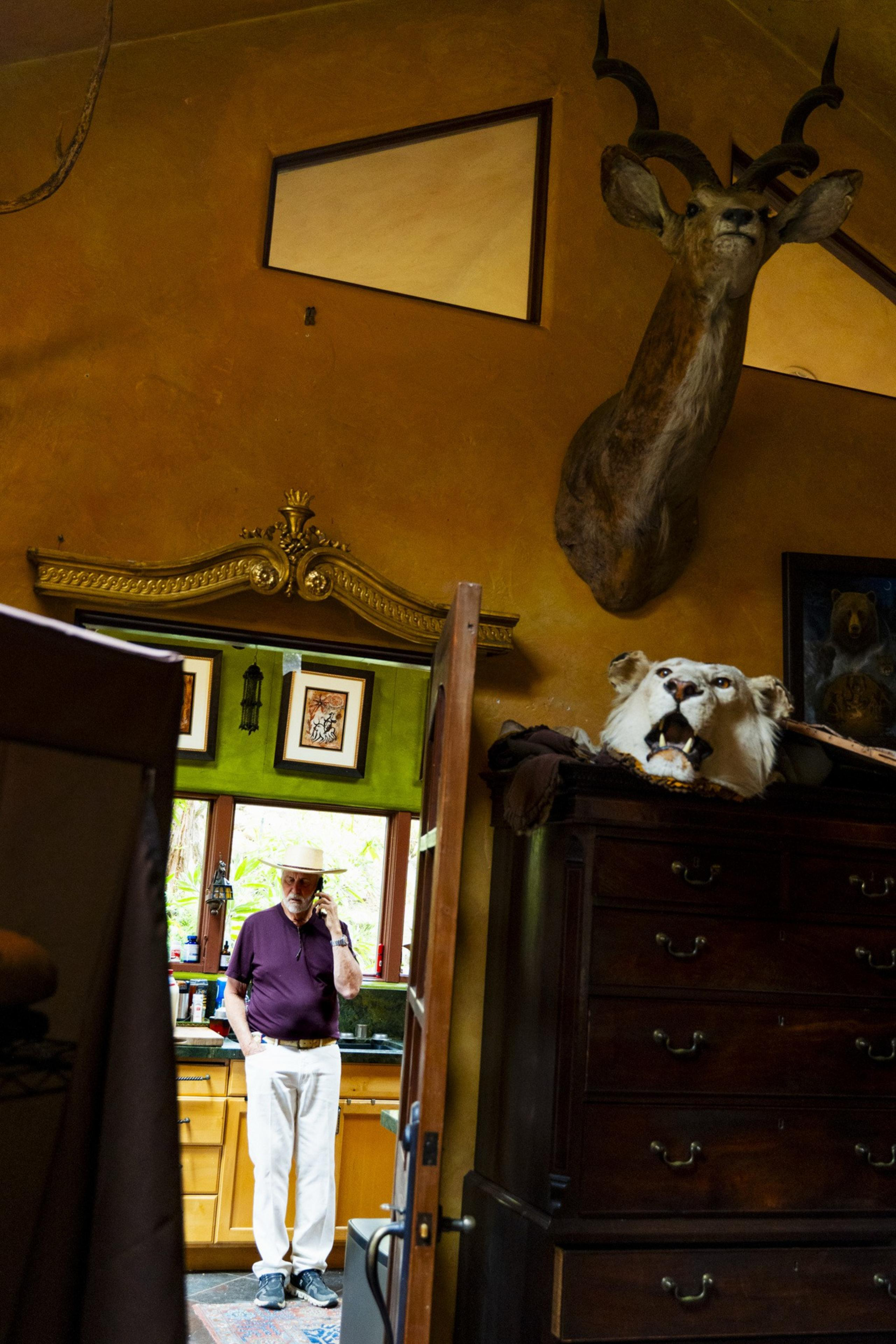 A man in a hat stands in a room with wooden cabinets, holding a phone. Mounted animal heads and a large wooden dresser are visible in the foreground.
