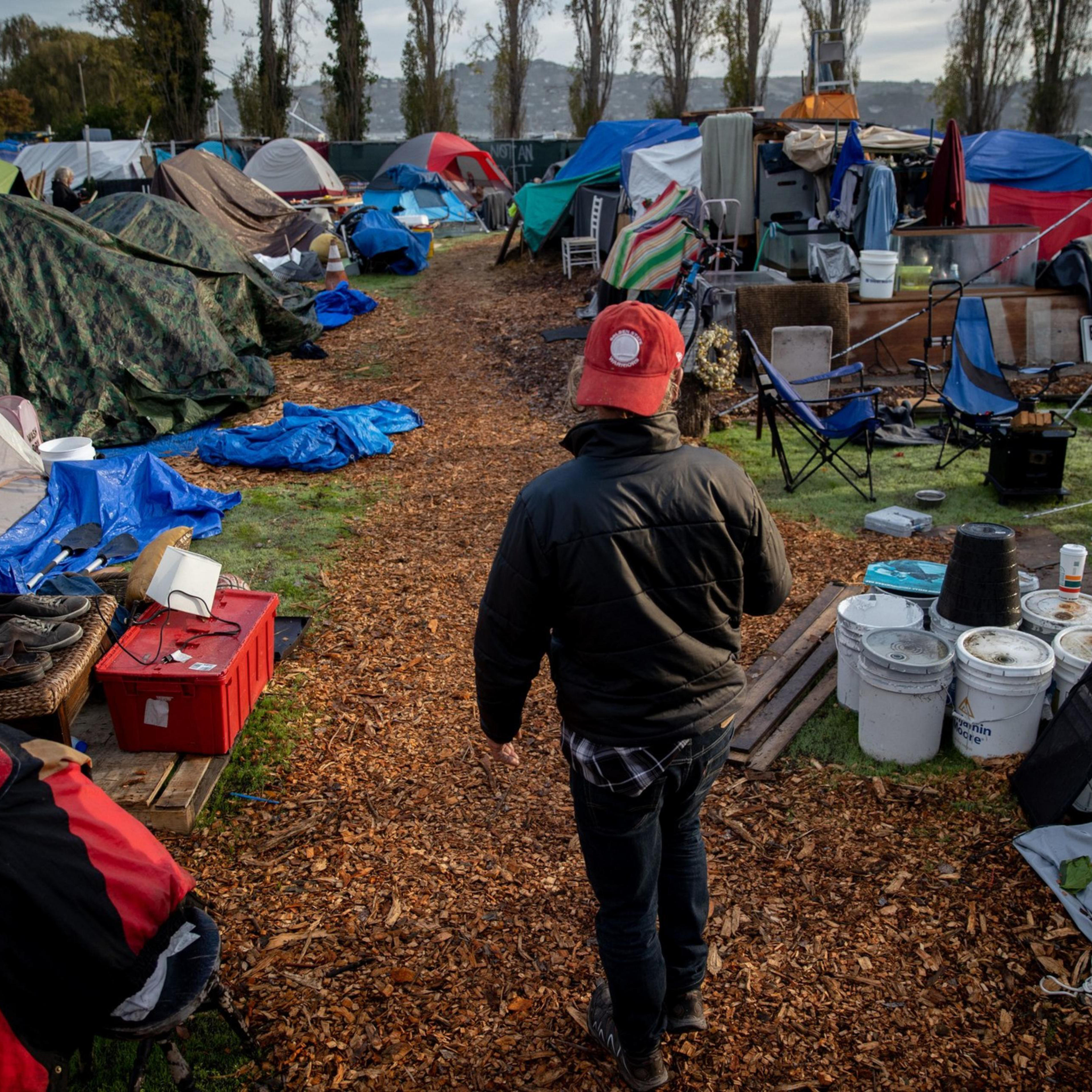 A person wearing a red cap and black jacket walks on a path through a crowded campsite with numerous tents, tarps, and scattered belongings on a grassy field.