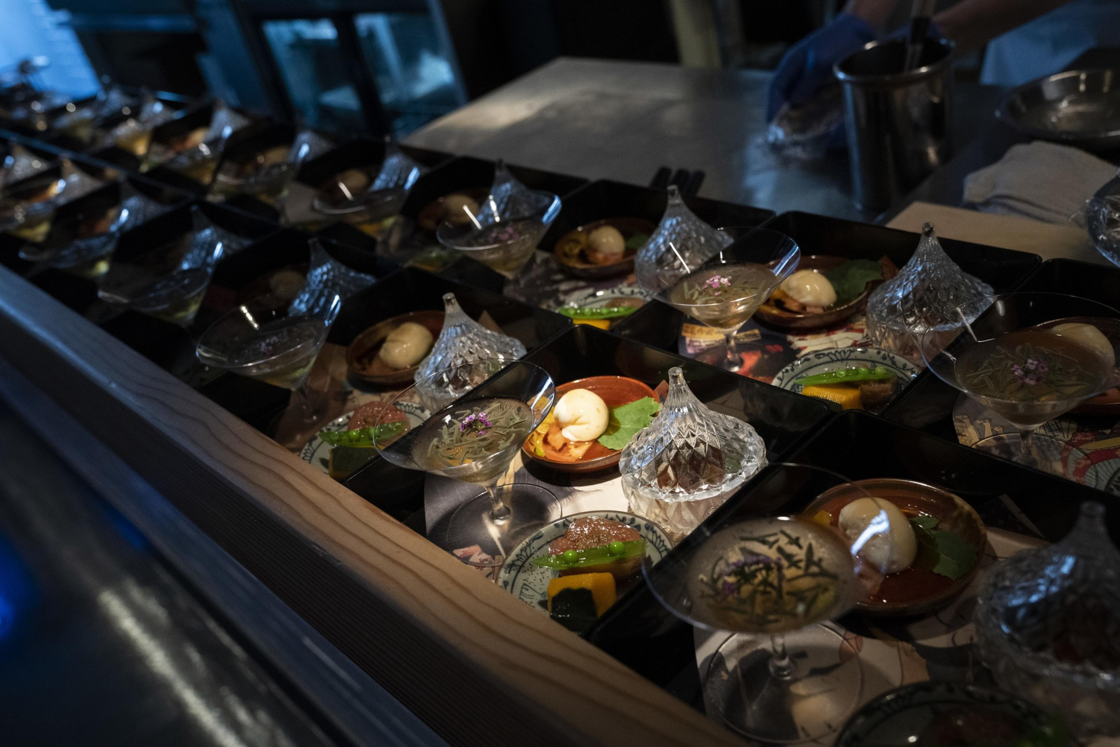 The image shows a row of elegant dishes on a wooden counter, featuring small glass bowls with intricate crystal lids and artfully presented appetizers.