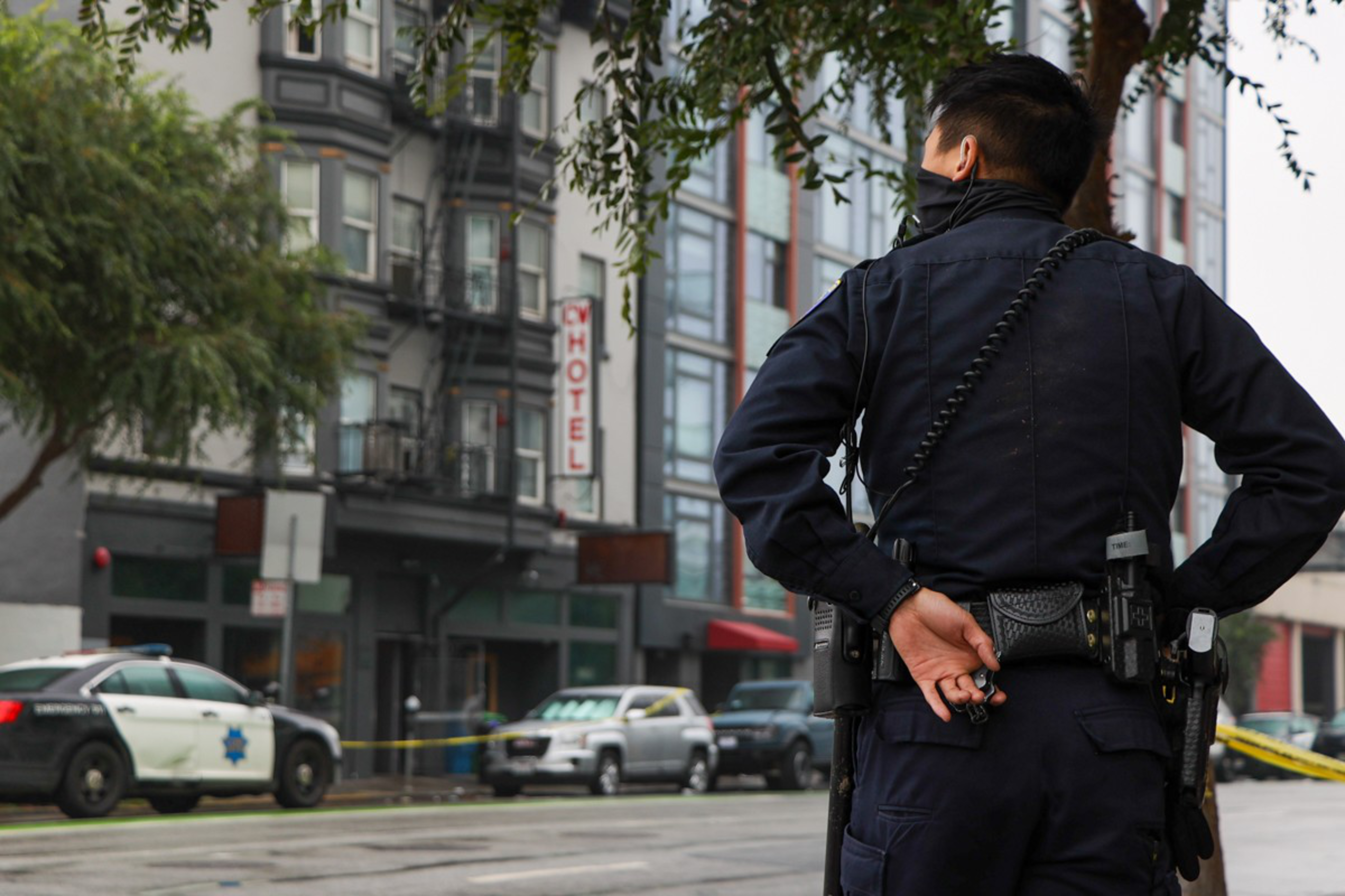 A police officer stands with hands behind back, facing a street blocked by police tape, near a hotel and several parked vehicles.