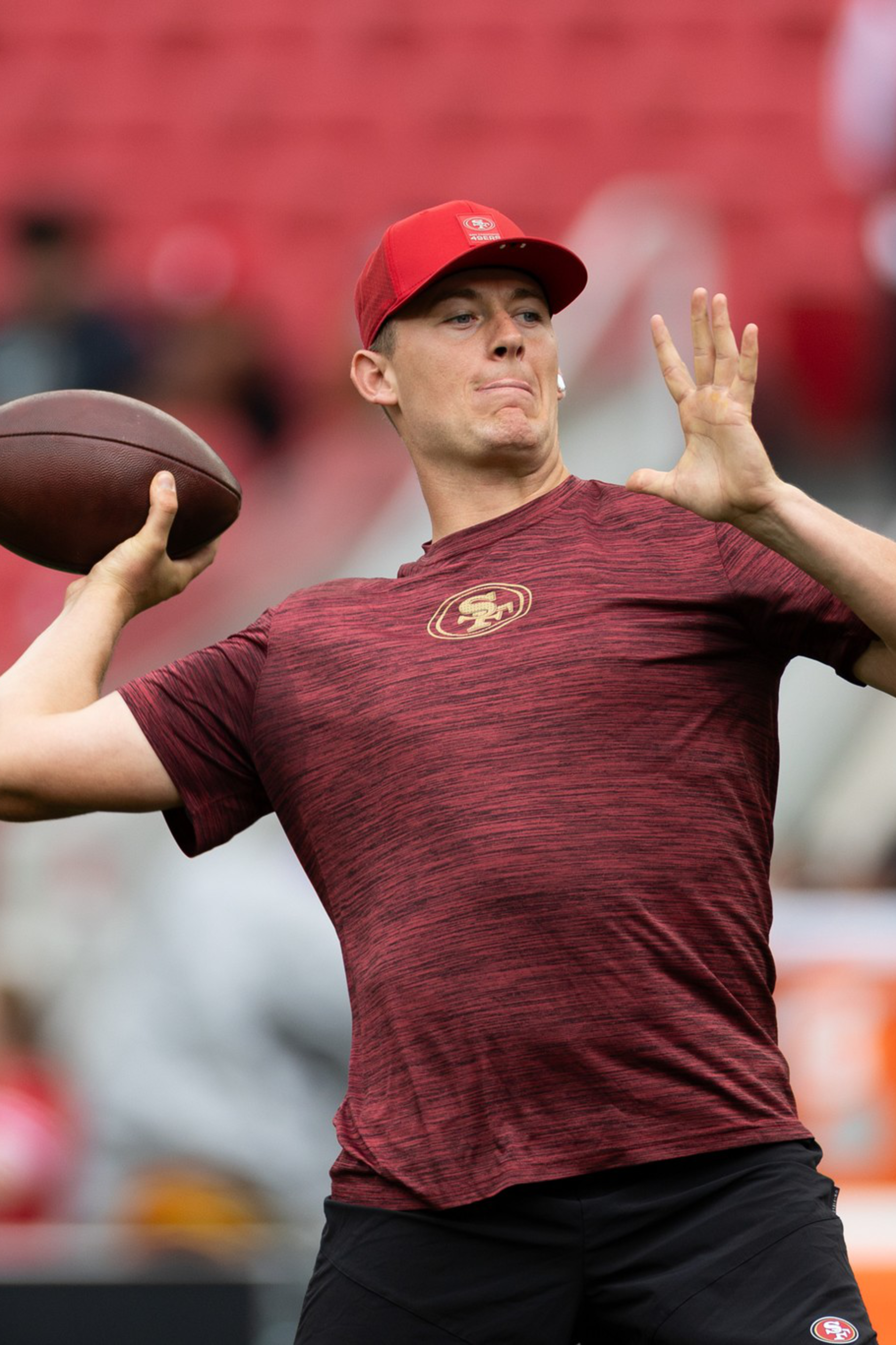 A football player in a red shirt and cap prepares to throw a football while another player in a similar outfit watches in a stadium.