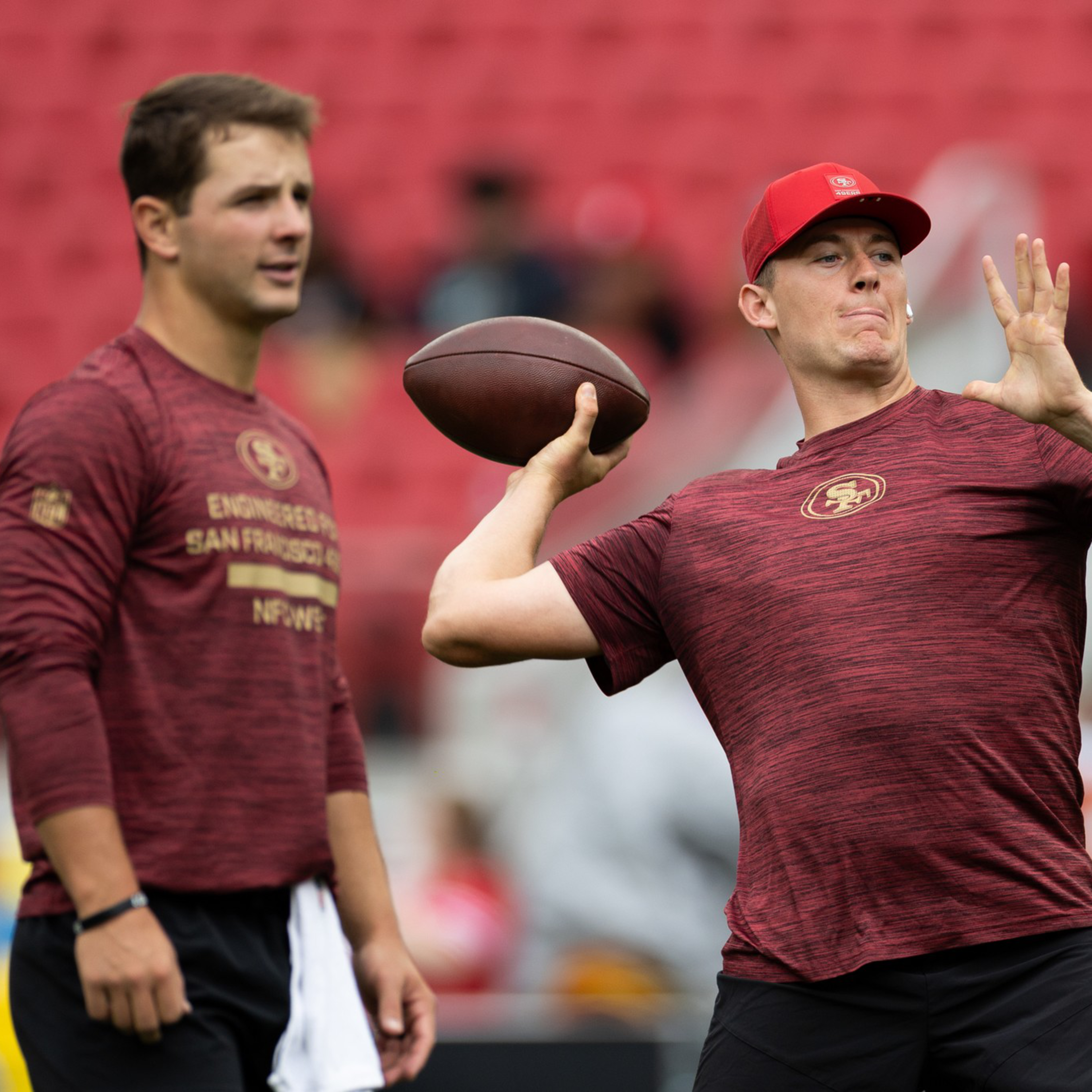 A football player in a red shirt and cap prepares to throw a football while another player in a similar outfit watches in a stadium.