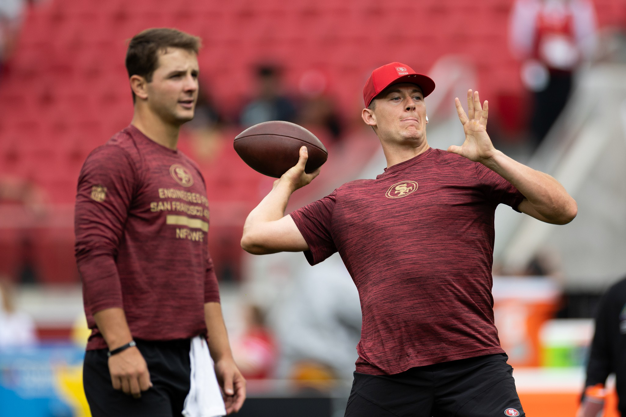 A football player in a red shirt and cap prepares to throw a football while another player in a similar outfit watches in a stadium.