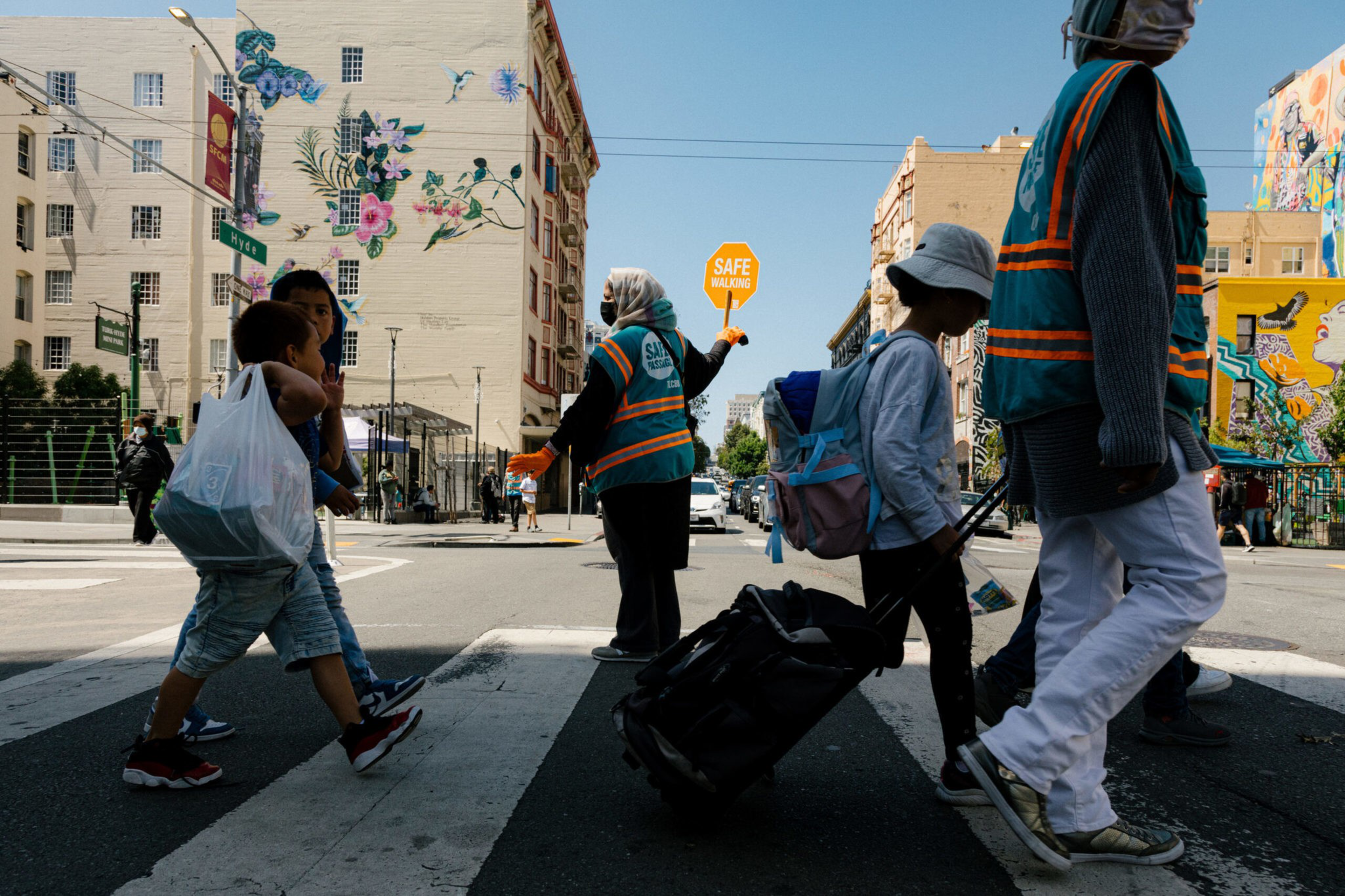A crossing guard holds a "SAFE WALKING" sign as pedestrians cross a sunny urban street with vivid murals.