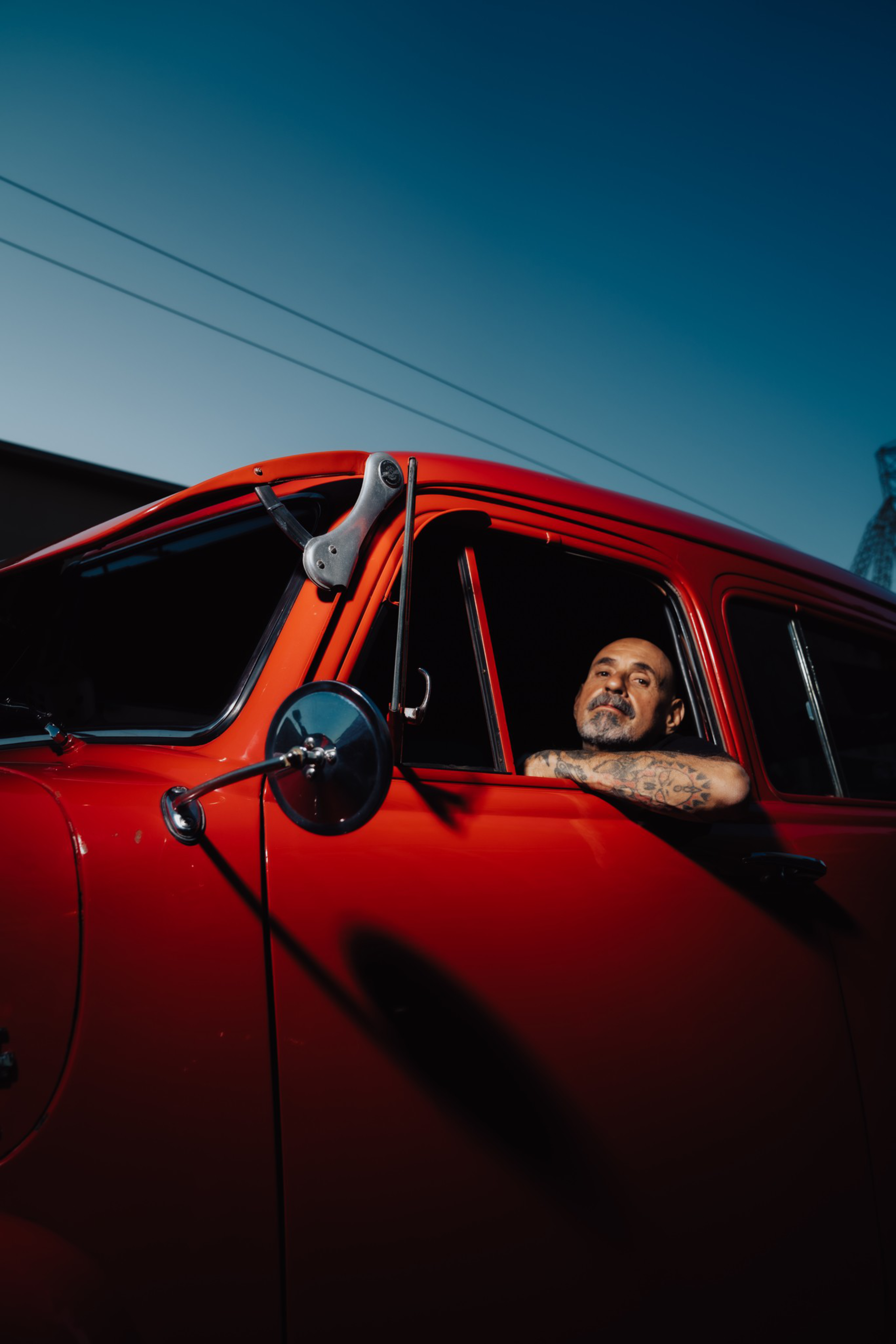 A man with tattoos rests his arm on the window of a vibrant red vintage truck against a deep blue sky at dusk.