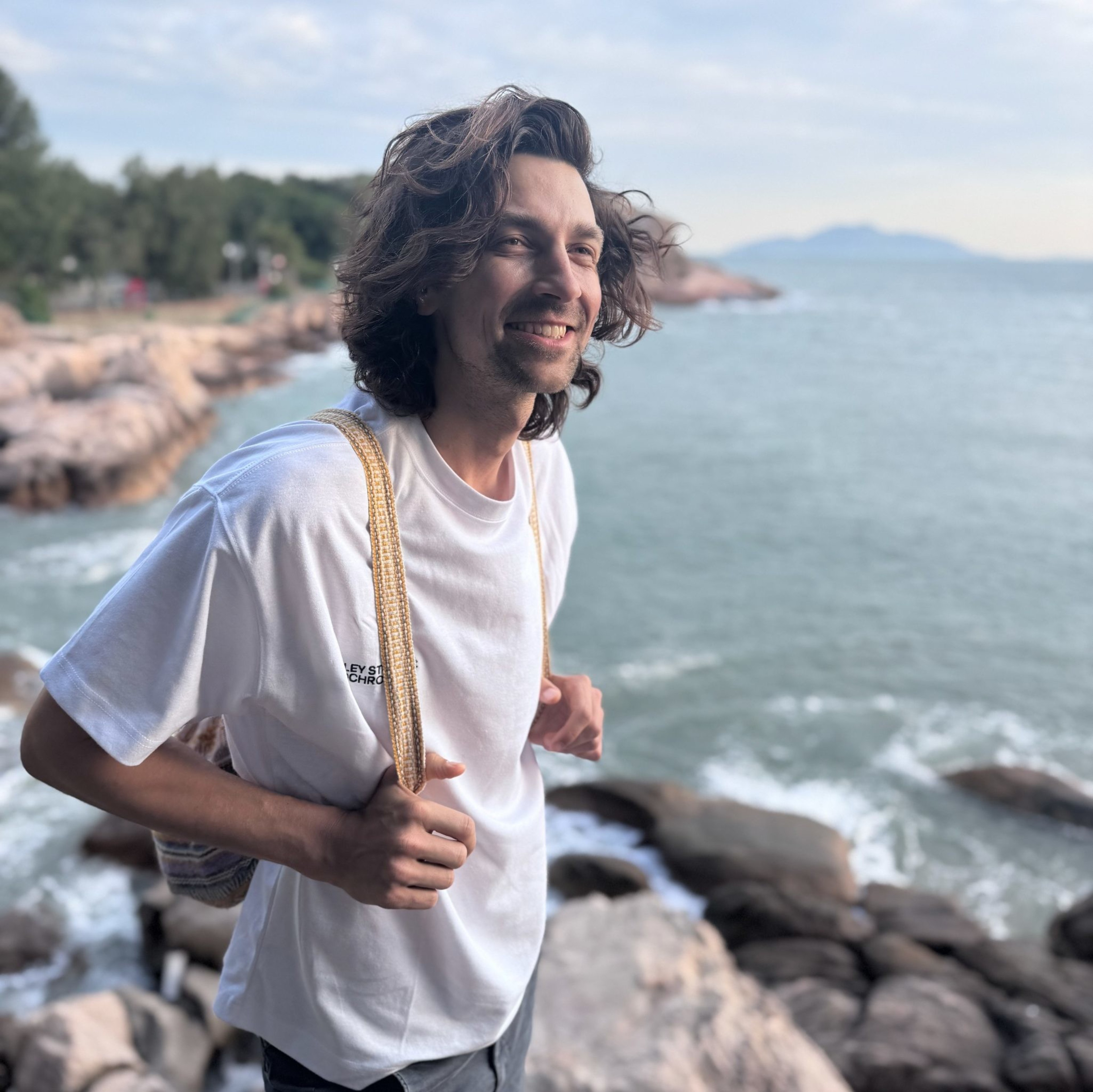 A person with wavy hair smiles while standing on rocky terrain by the sea, wearing a white shirt and carrying a woven bag. The sky is partly cloudy.