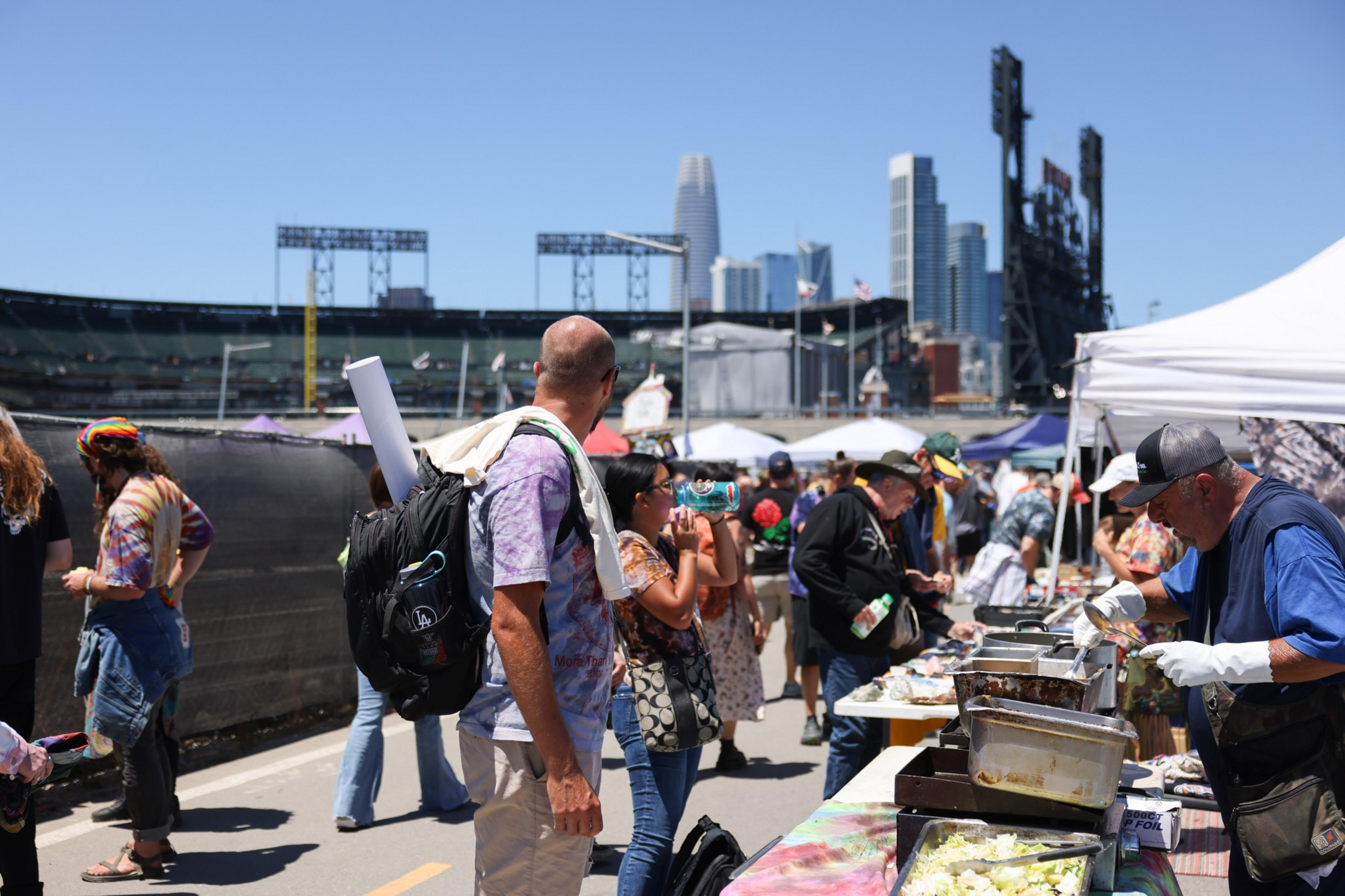 People browse food stalls at an outdoor market near a stadium with city skyscrapers visible in the background on a sunny day.