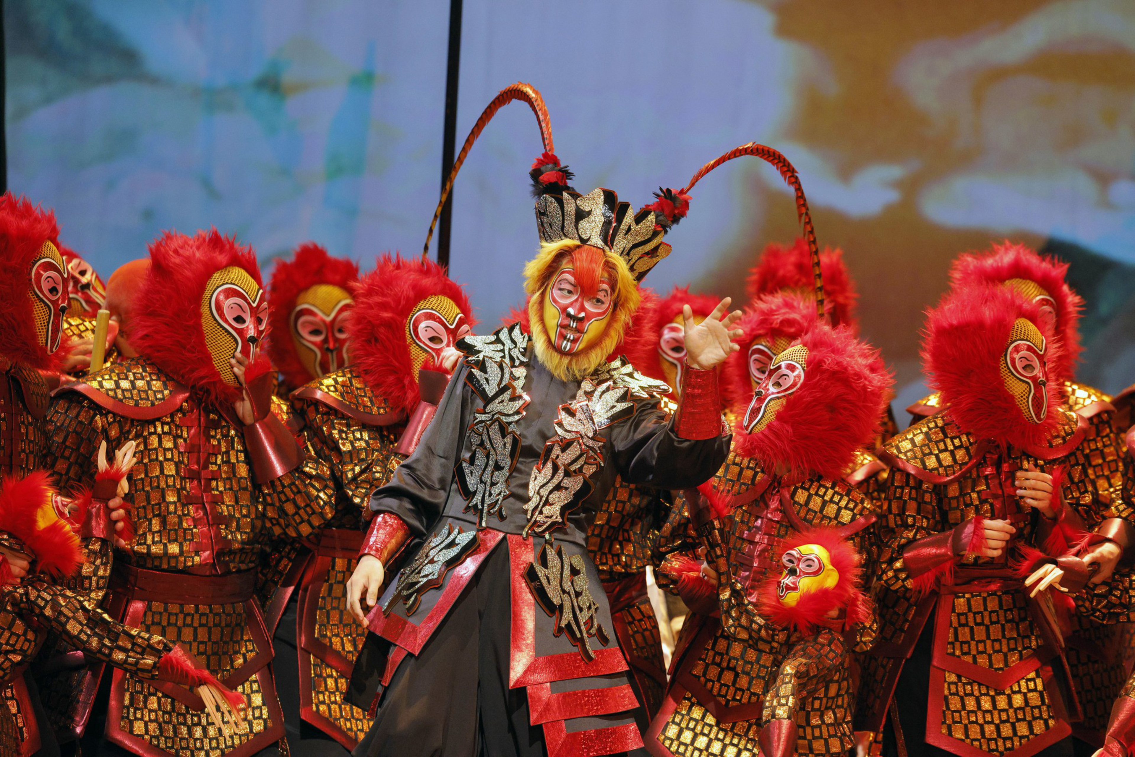Performers wear elaborate costumes with red monkey masks and gold patterned armor, with one central figure in a detailed black and red outfit with long antennae.