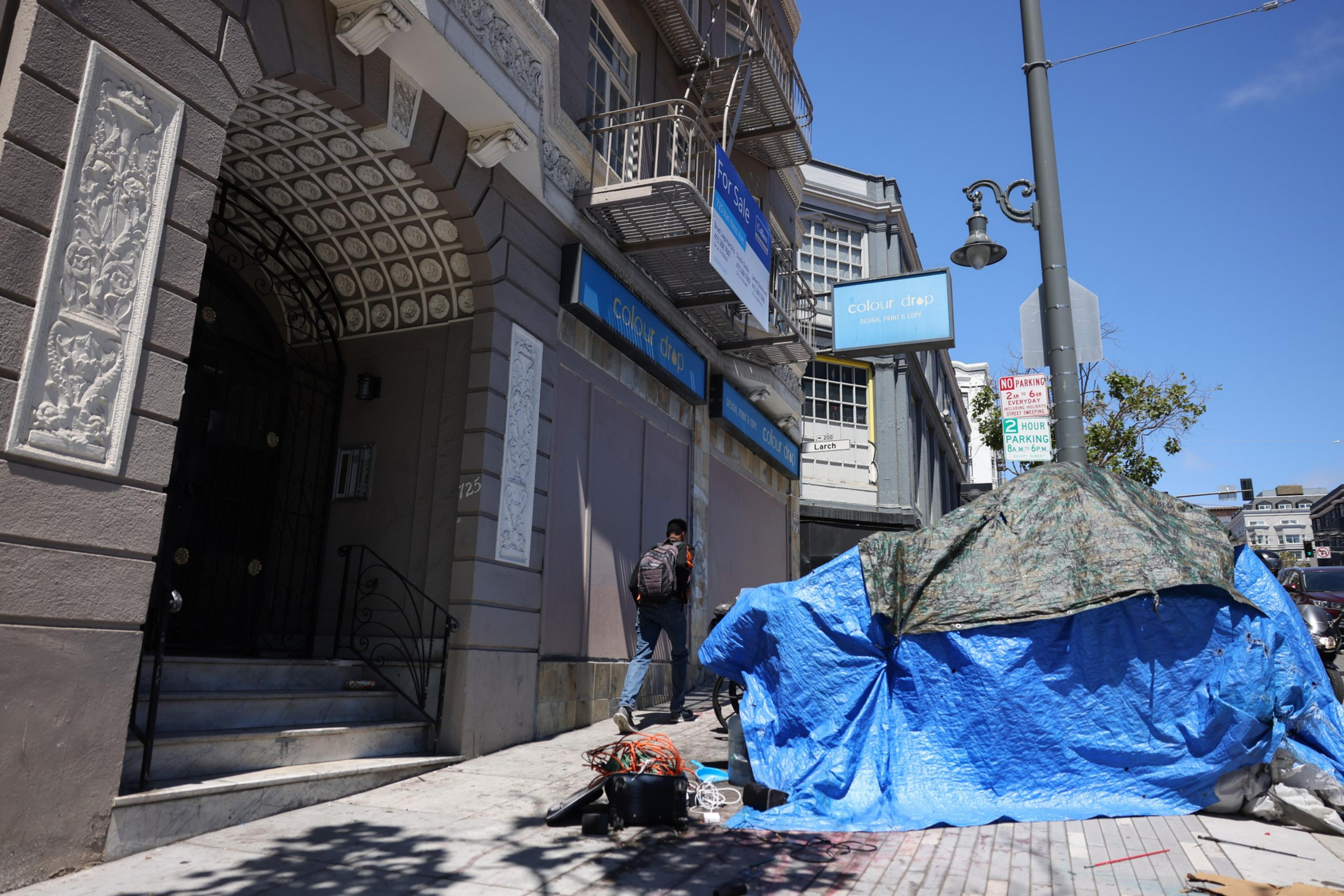 A man with a backpack walks past a building with decorative stonework, while a large tarp-covered structure occupies the sidewalk near a streetlamp.