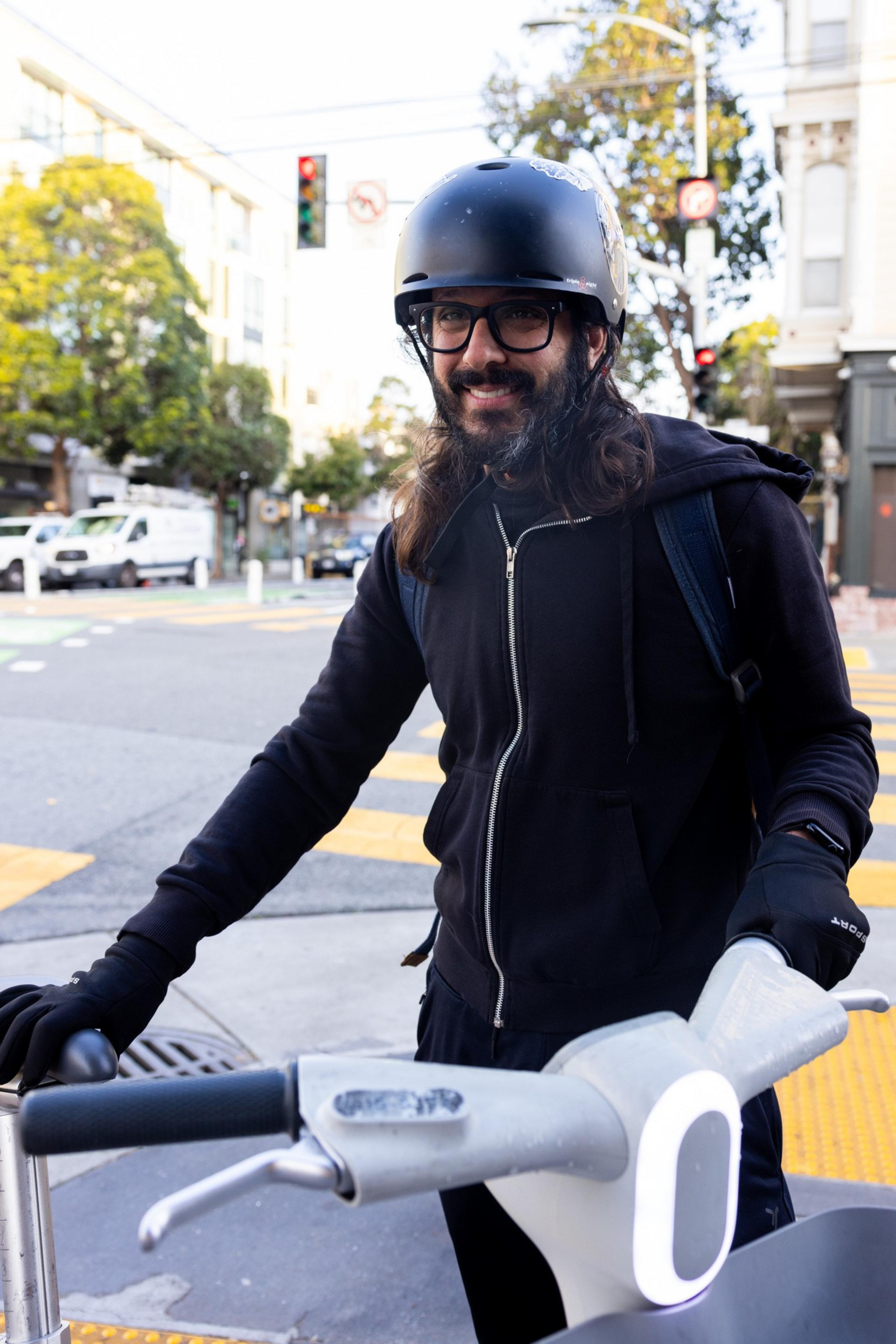 A person with long hair and glasses wears a black helmet and jacket, standing on a city street with a scooter. Trees and buildings are in the background.