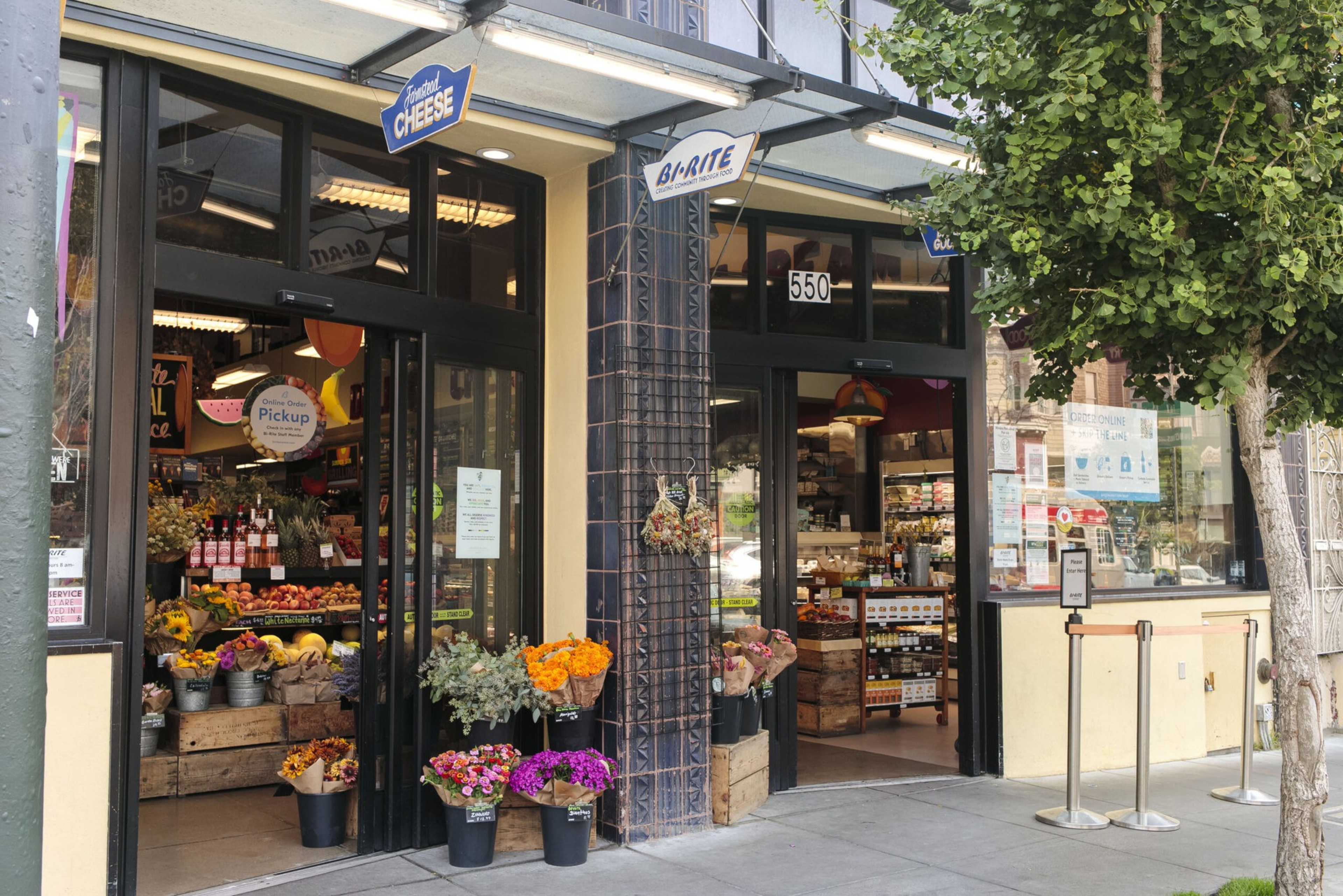 A grocery store entrance displays colorful flower bunches outside, with fresh produce, wine bottles, and signage visible inside.