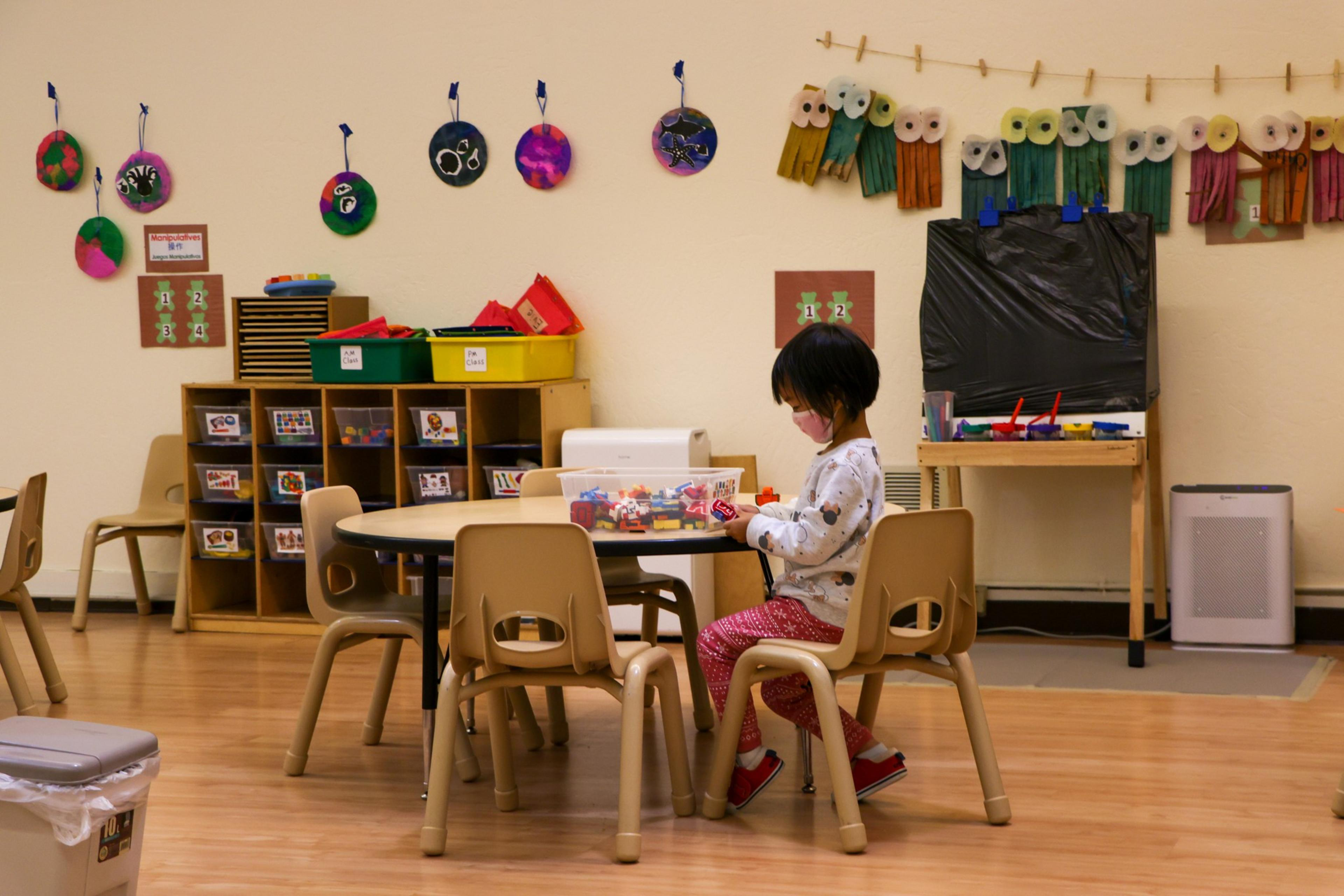A child sits at a small table in a classroom filled with colorful art supplies and decorations on the walls, engaging with toys.