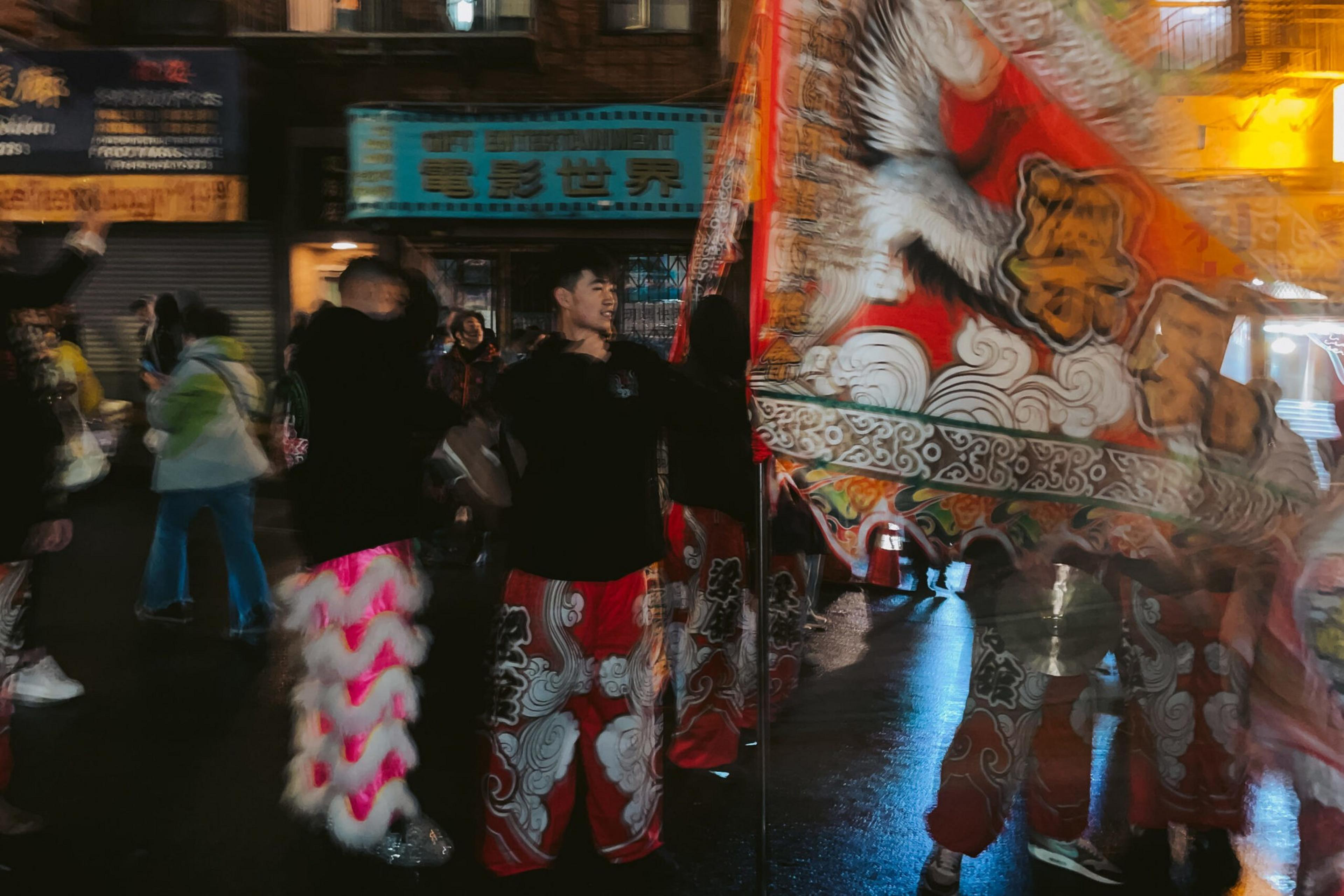 People in colorful traditional clothing perform a dragon or lion dance on a wet street at night, surrounded by illuminated signs and onlookers.