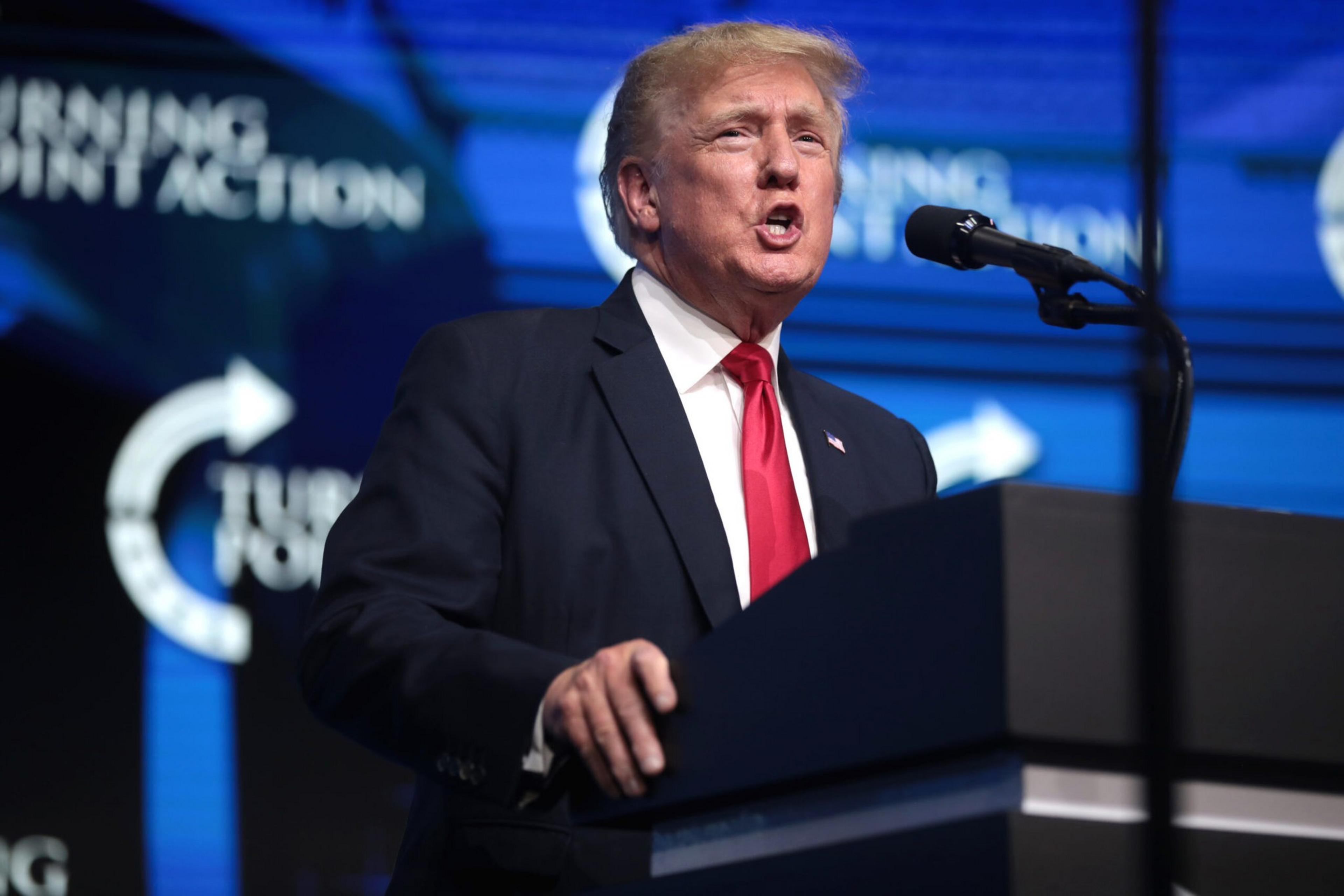 A man in a dark suit with a red tie speaks passionately at a podium with a microphone against a blue backdrop featuring the words &quot;Turning Point Action.&quot;